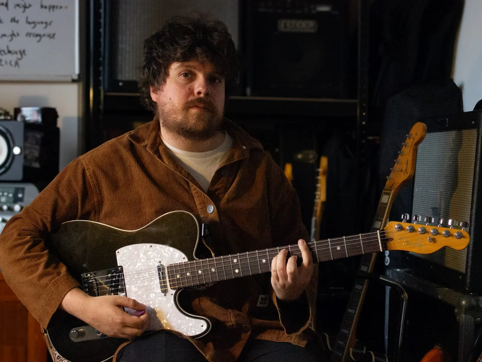 A man with curly hair and a beard sitting in a music studio, holding a black Fender electric guitar. The room has musical equipment, amplifiers, and guitars in the background.