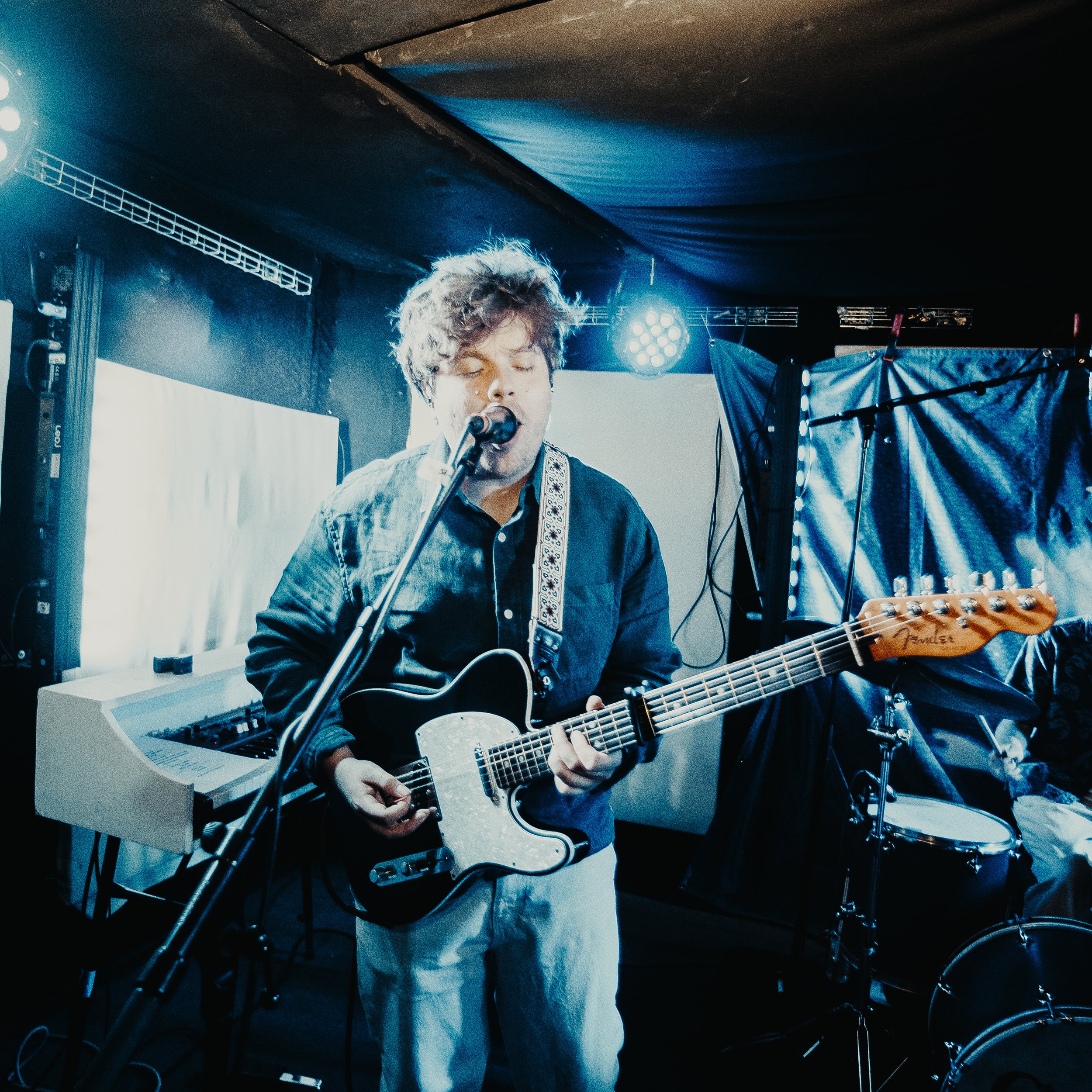 A young man with grayish, curly hair playing an electric guitar and singing into a microphone during a live music performance, with stage lights and equipment in the background.