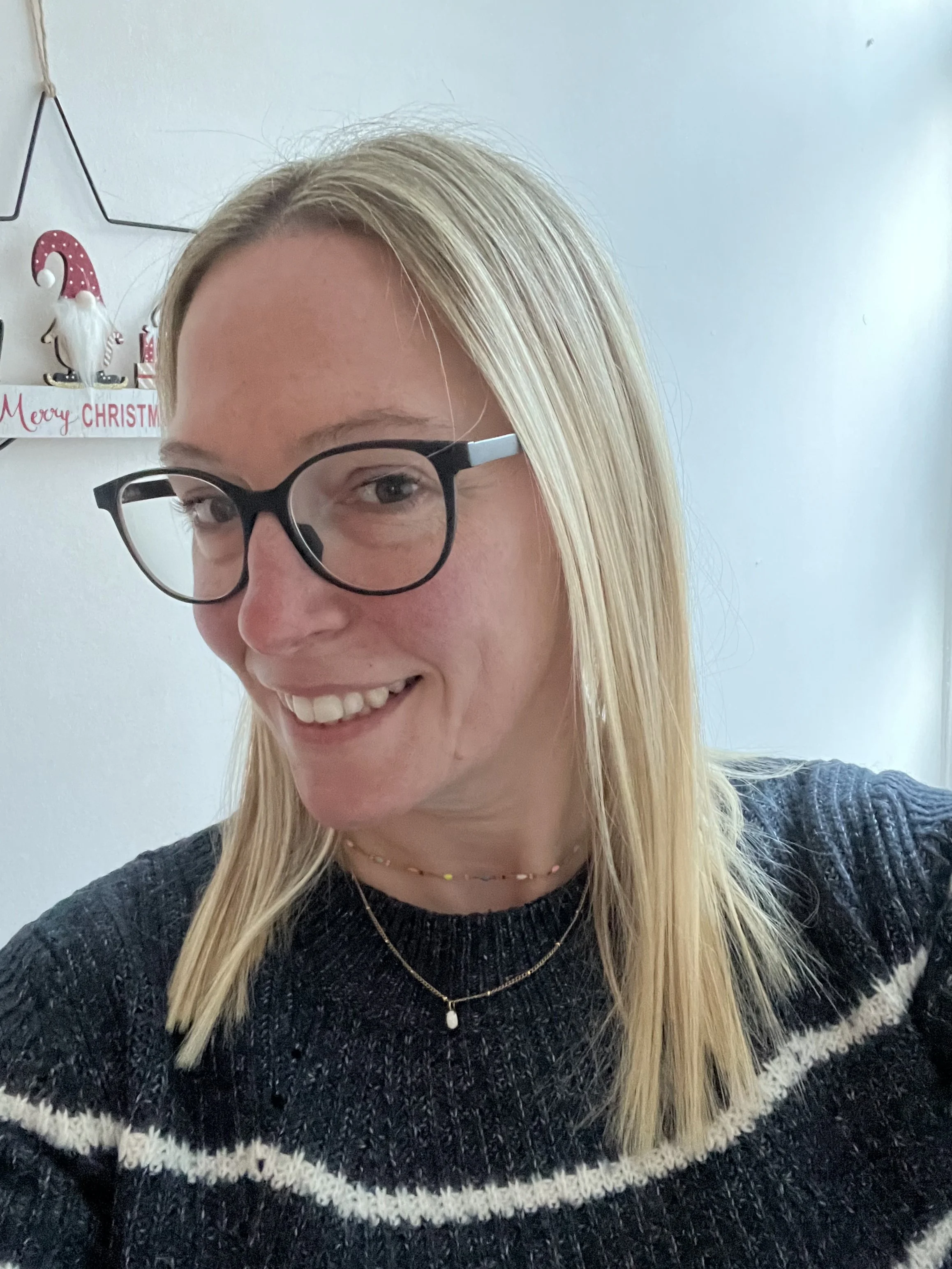Close-up of a smiling woman with blonde hair, wearing glasses, a necklace, and a gray and white patterned sweater, indoors with Christmas decorations in the background.