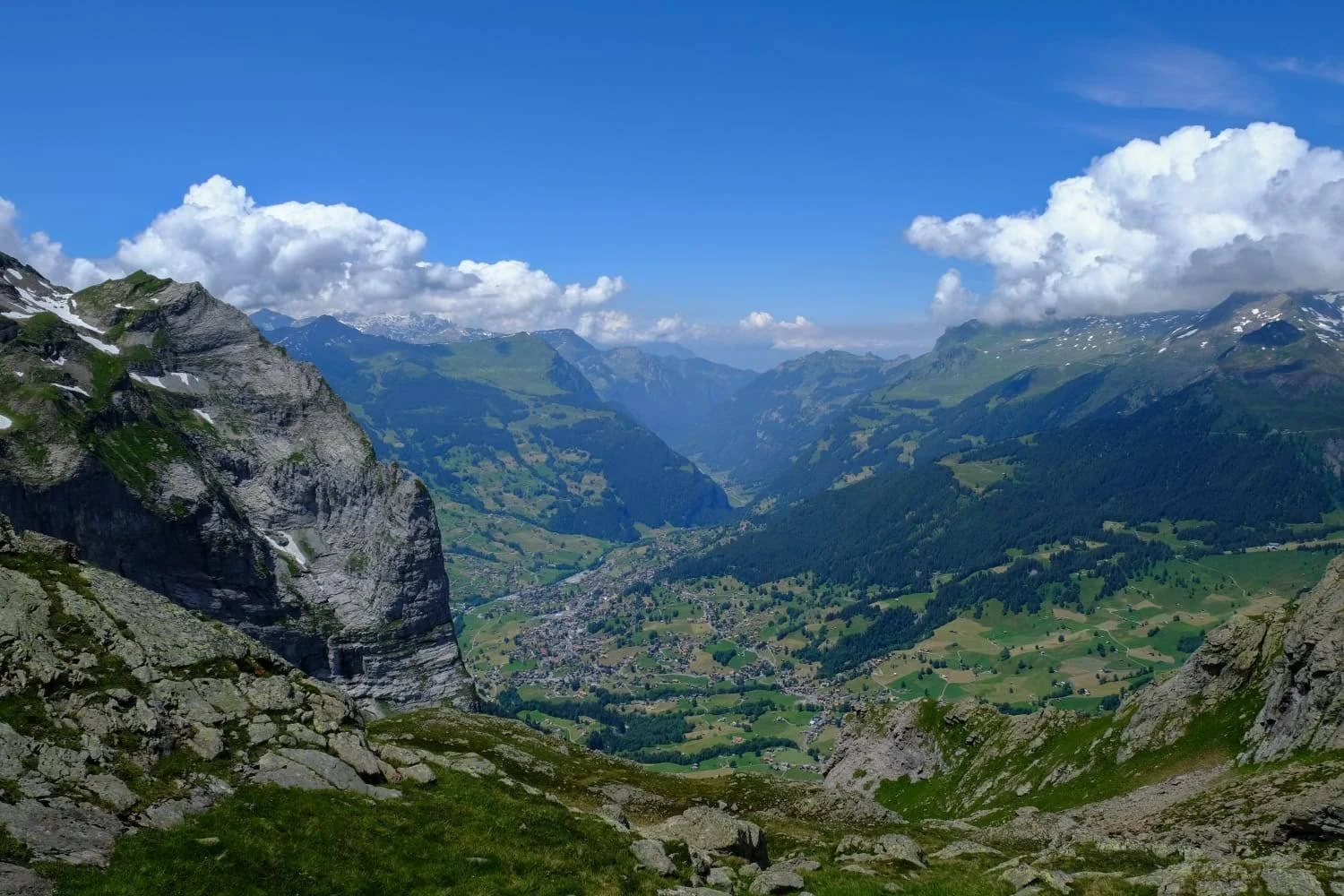 A panoramic view of a lush green valley surrounded by towering mountains with patches of snow, under a partly cloudy blue sky.