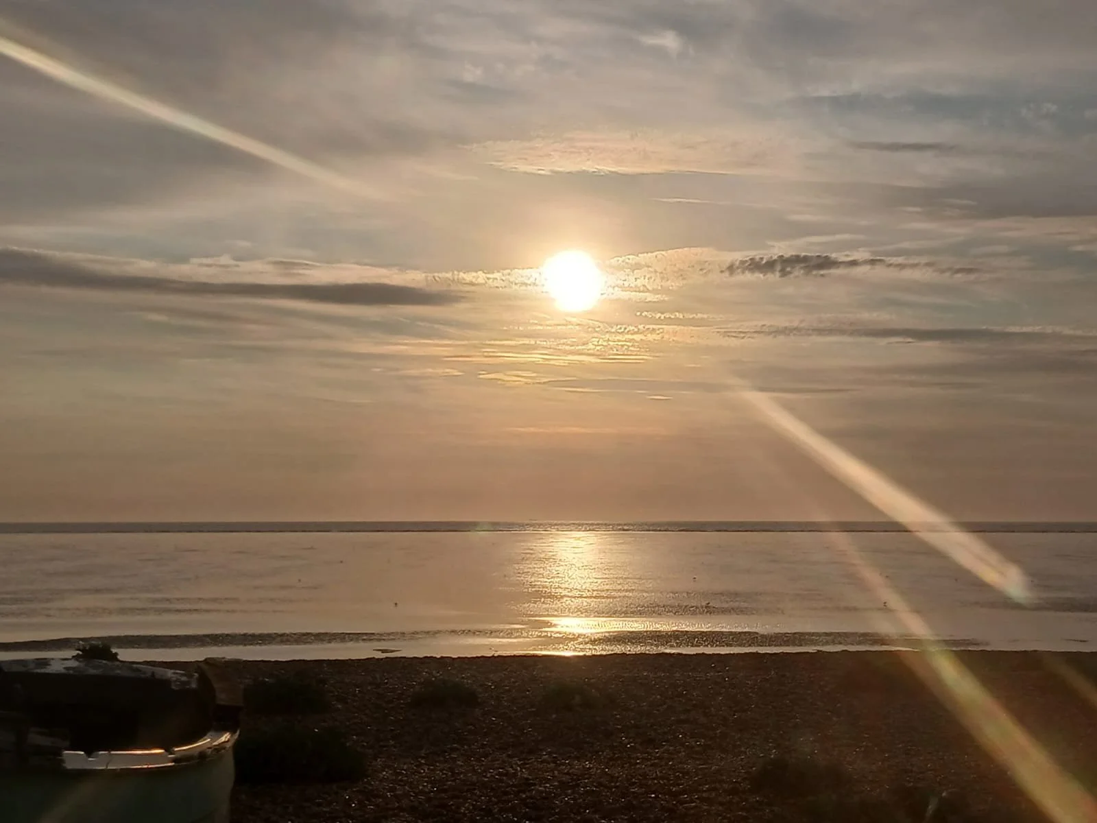 Sunset over the ocean with clouds in the sky and rays of sunlight reflecting on the water, viewed from a shoreline with a rocky beach.