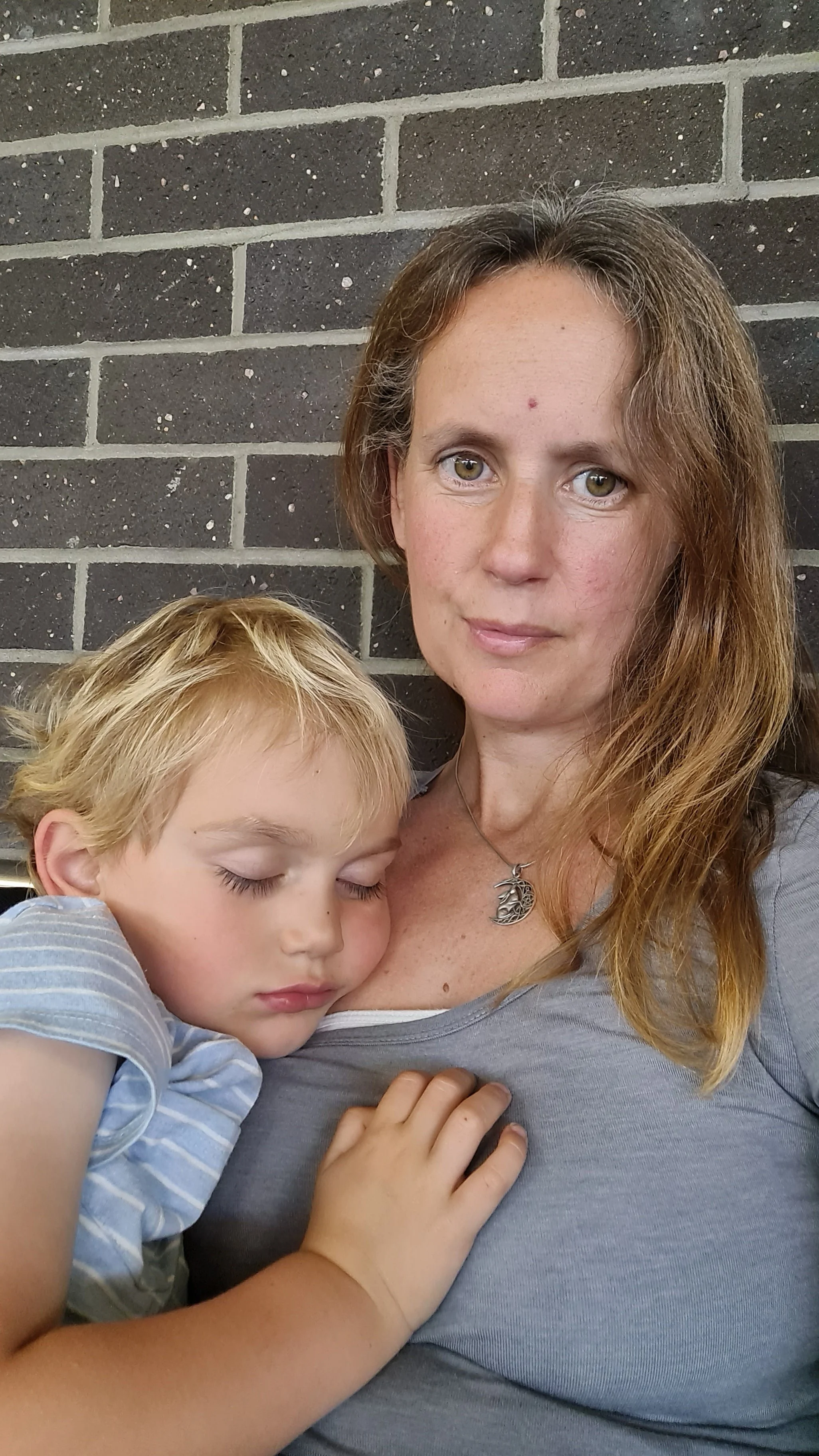 A woman with long, wavy, reddish-brown hair and a silver necklace sits against a black brick wall, holding a young boy with blonde hair and light skin, who is sleeping with his head resting on her chest.