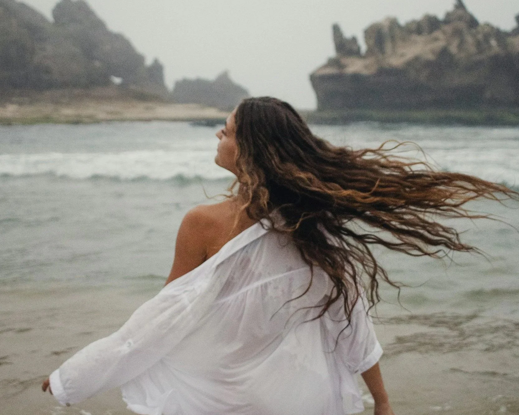 A woman with long, wavy hair standing on a beach, wrapped in a white fabric, looking towards the ocean with rocky cliffs in the background.