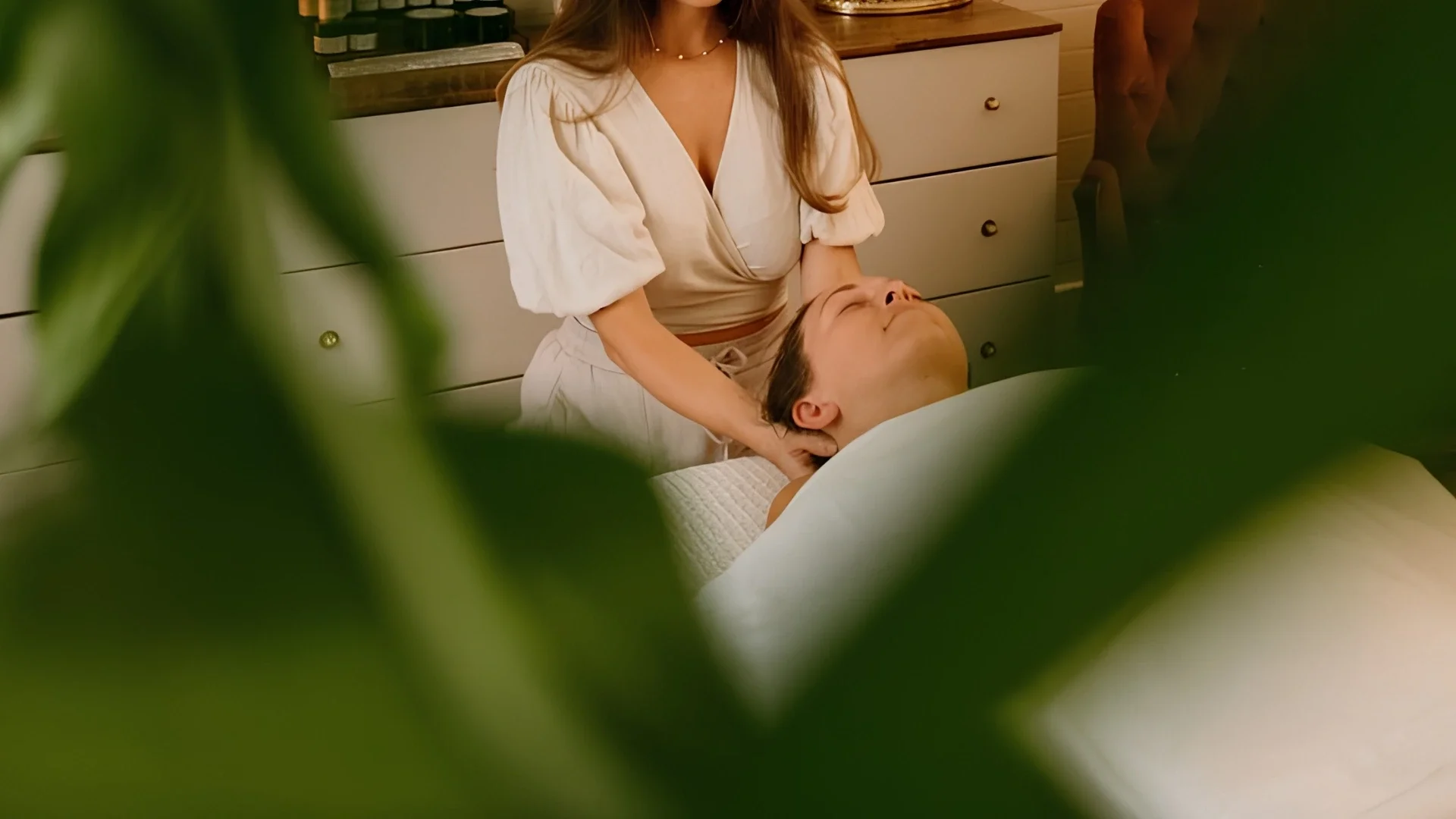 A woman giving a massage to a man lying on a massage table, seen through green leaves, in a cozy, wood-paneled room.