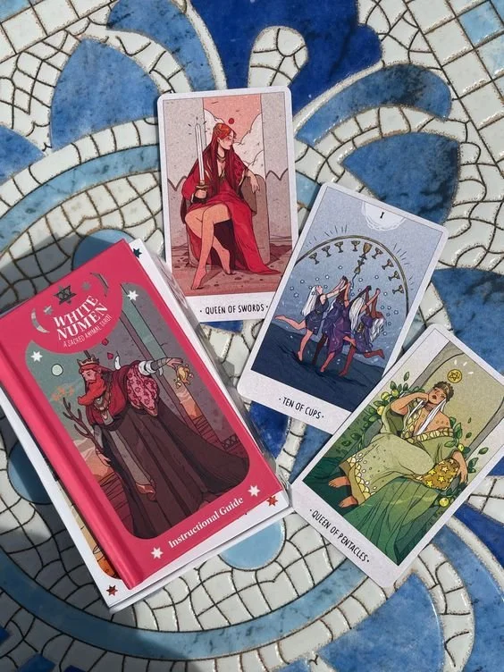 A set of tarot cards and a guidebook on a mosaic surface. The tarot cards include Queen of Swords, Ten of Cups, and Queen of Pentacles. The guidebook is titled "White Numbet" and shows a cartoon witch holding a wand.
