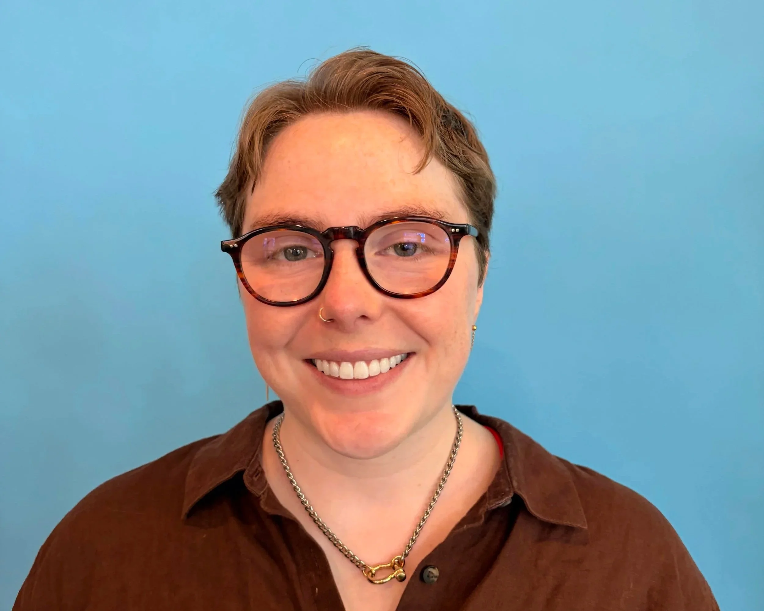 Close-up of a smiling woman with short hair, wearing glasses, a nose ring, a brown shirt, and a silver chain necklace, standing against a blue background.