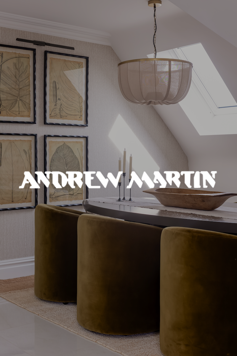 Modern dining room with a round table, green velvet chairs, a sculptural wooden bowl, candelabra with three candles, framed botanical prints on the wall, a ceiling light fixture, and a skylight in a bright space.