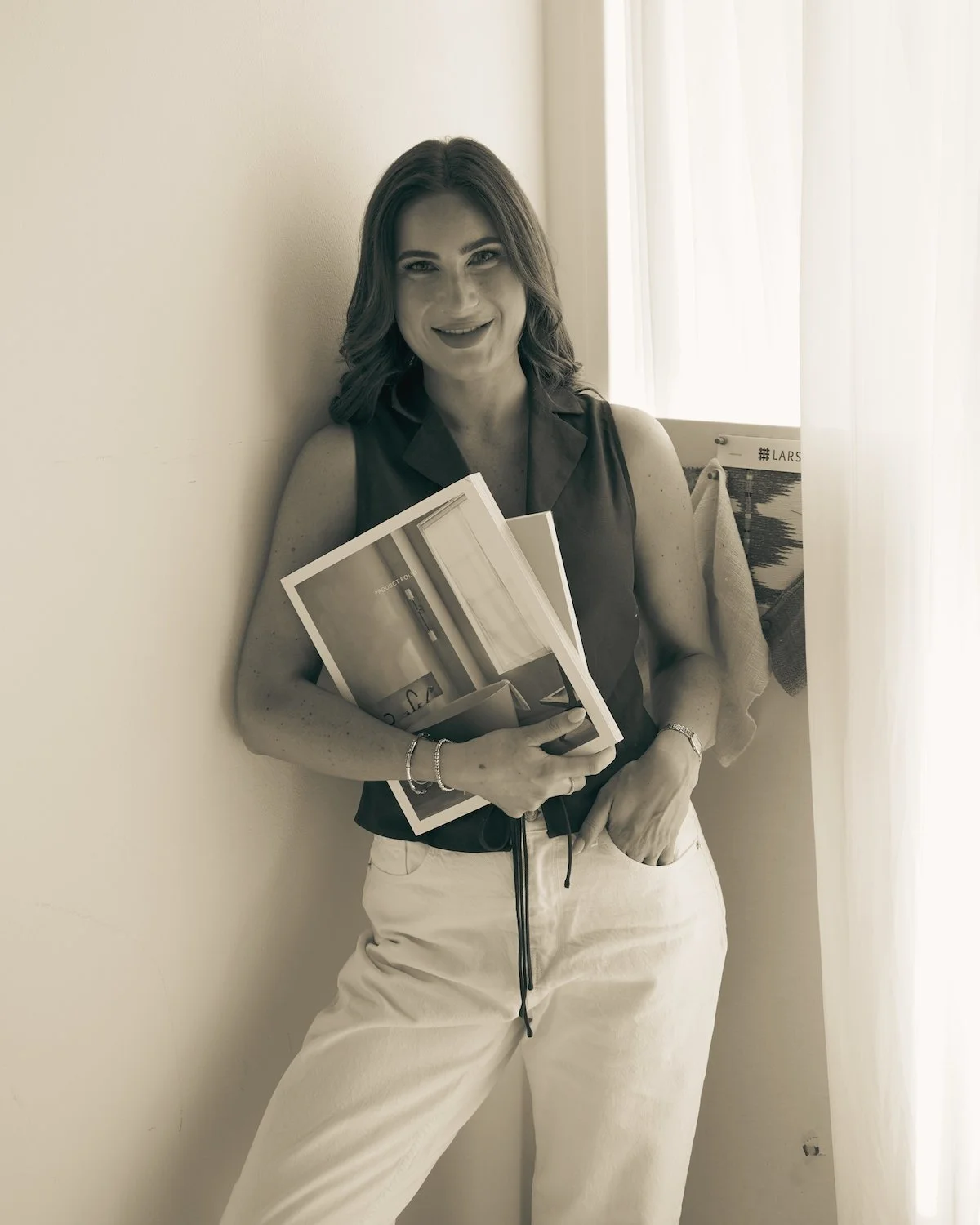 Black and white photo of a smiling woman with shoulder-length hair leaning against a wall, with arms crossed, wearing a sleeveless top and light-colored pants. There is a curtain and a magazine or brochure with visible text in the background.