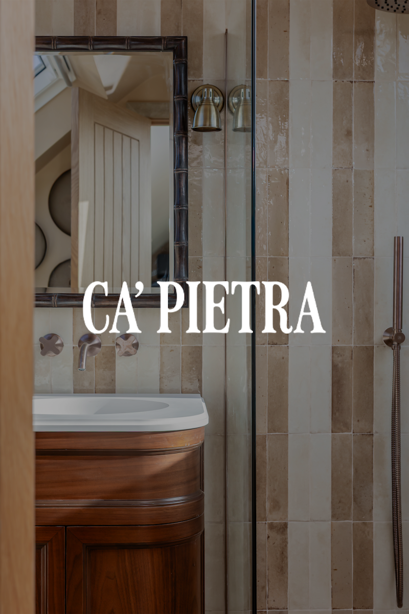 Bathroom with a wooden vanity with a white sink, a mirror, two brass wall sconces, and beige tiled walls and shower area.