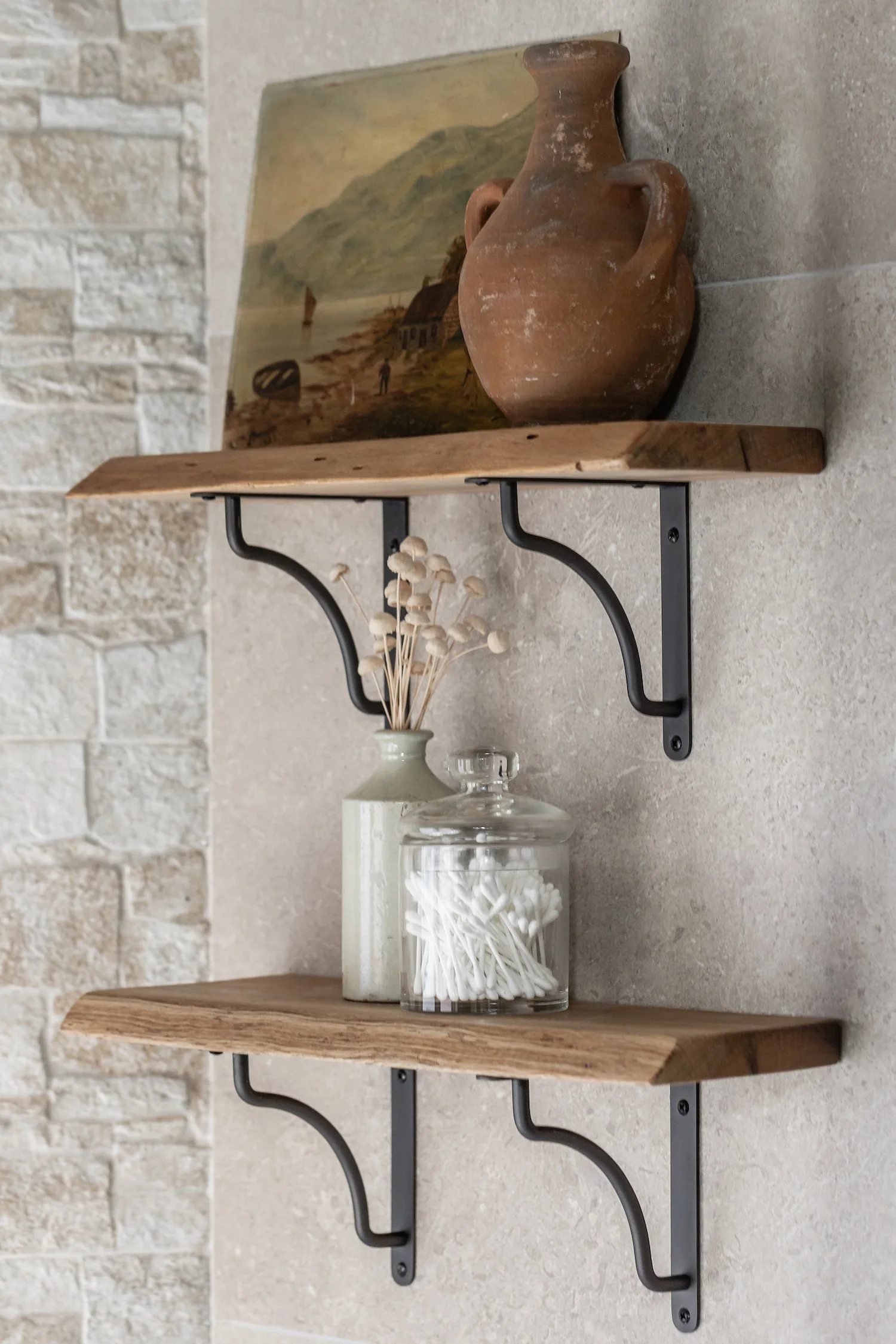 Two wall-mounted wooden shelves supported by black metal brackets, with decorative items including a glass jar filled with white cotton swabs, a white ceramic vase with dried white mushrooms, and a framed landscape painting of a coastal scene with hills and houses.