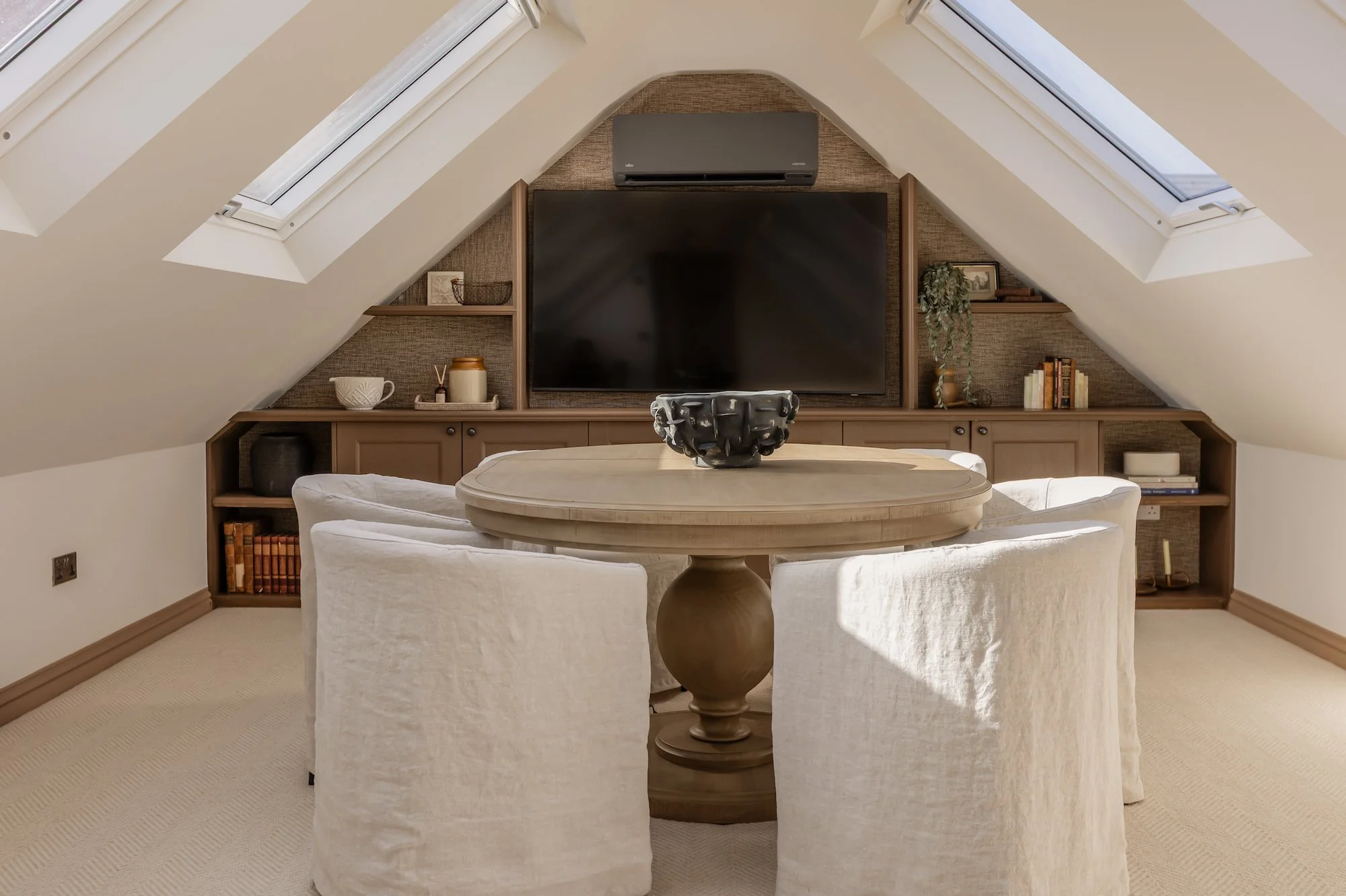 A cozy attic living space featuring a round wooden table surrounded by four cream-colored chairs, a flat-screen TV mounted on a wood-paneled wall, and two skylight windows providing natural light.