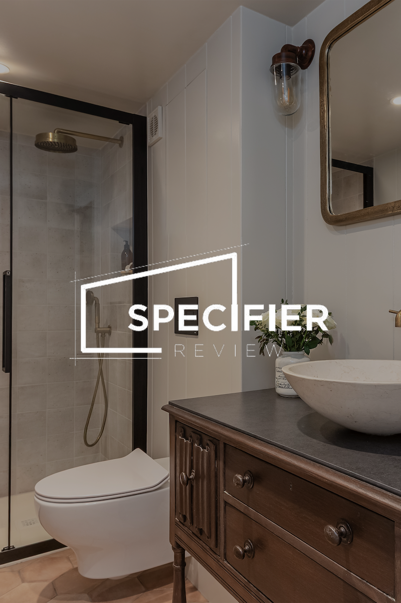 A modern bathroom featuring a shower with a brass rain showerhead, a white toilet, a vanity with a vessel sink, a mirror with a wooden frame, a small plant, and a wall-mounted light fixture.