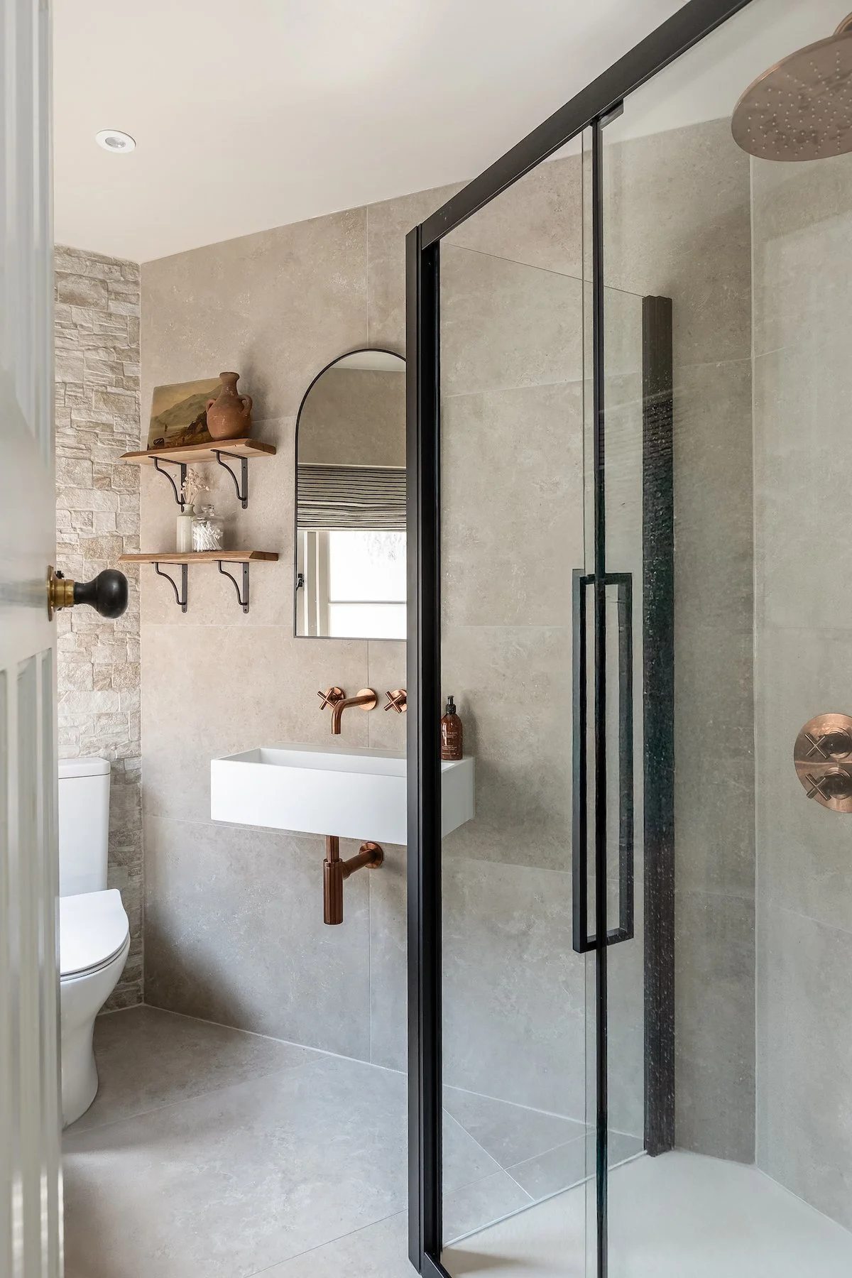 Modern bathroom with beige tiled walls, a black-framed glass shower, a wall-mounted sink with copper fixtures, a mirror, and a decorative shelf with vases and a painting.