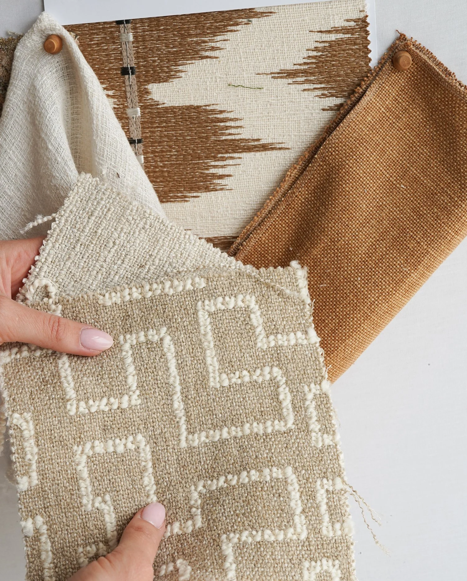 Close-up of woven textiles, including a beige and white textured fabric being held, a patterned cream and beige rug, and a brown textured rug, arranged on a white surface.