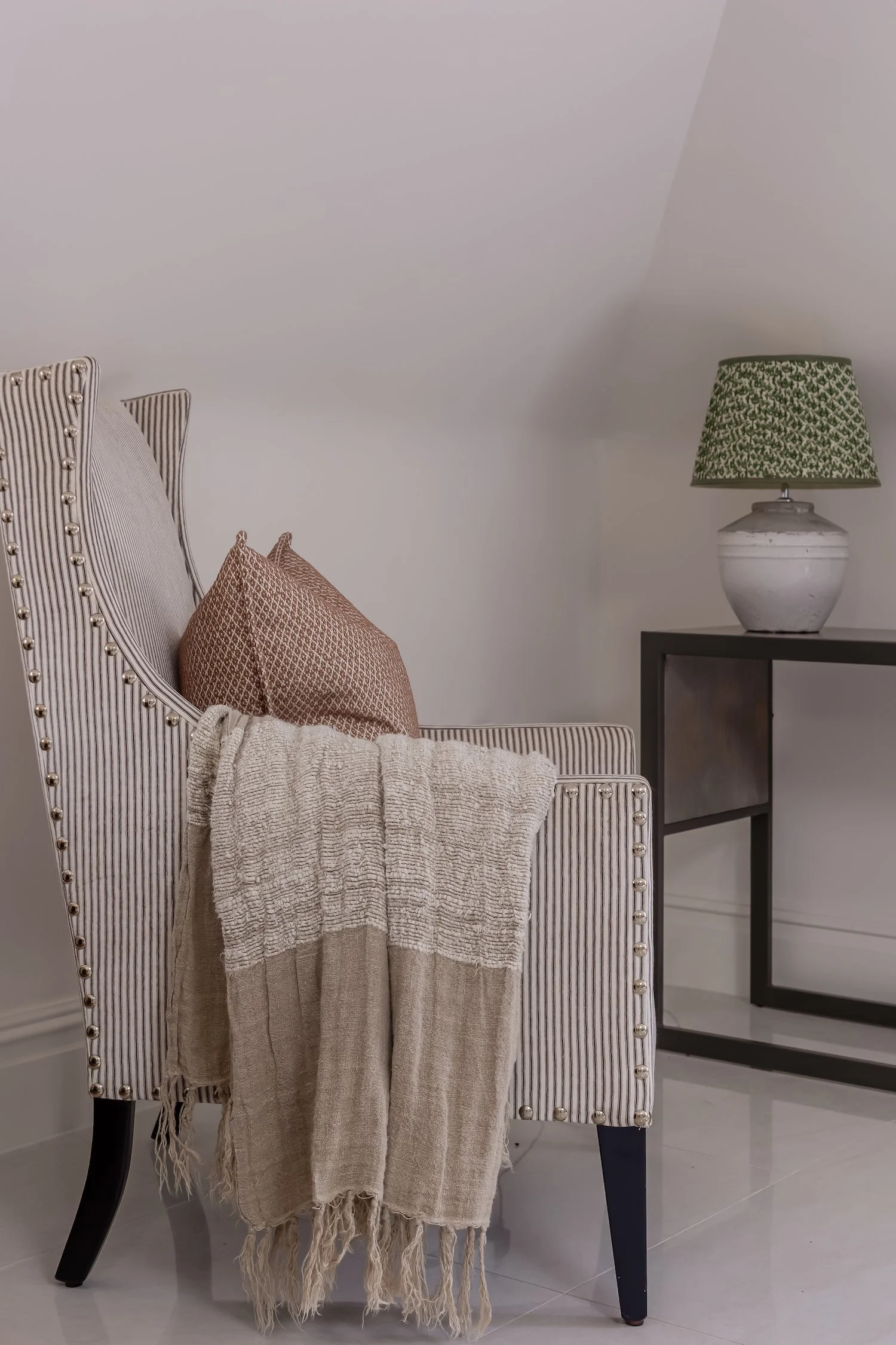 A beige and black striped upholstered chair with dark wooden legs, draped with a beige woven throw and a brown patterned pillow. To the right, a black side table with a white ceramic lamp featuring a green patterned shade is visible. The room has white walls and light-colored tile flooring.