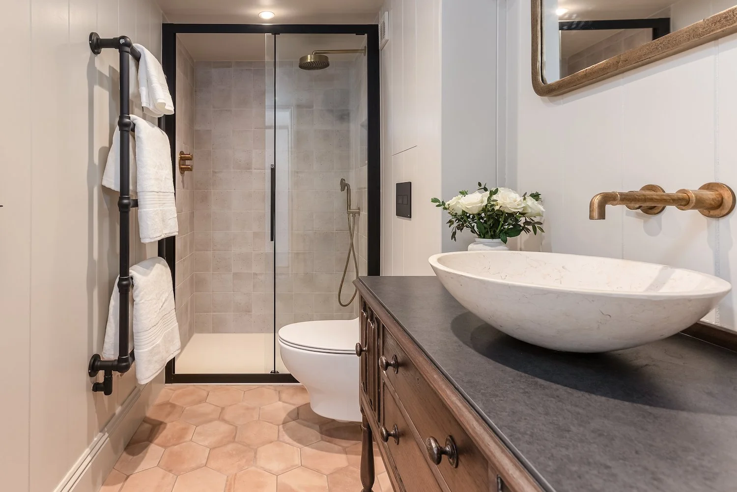 A modern bathroom featuring a walk-in shower with a glass door, a white toilet, a wooden vanity with a vessel sink, a gold-colored faucet, a mirror, and a towel rack with white towels. There is a vase with white flowers on the vanity.