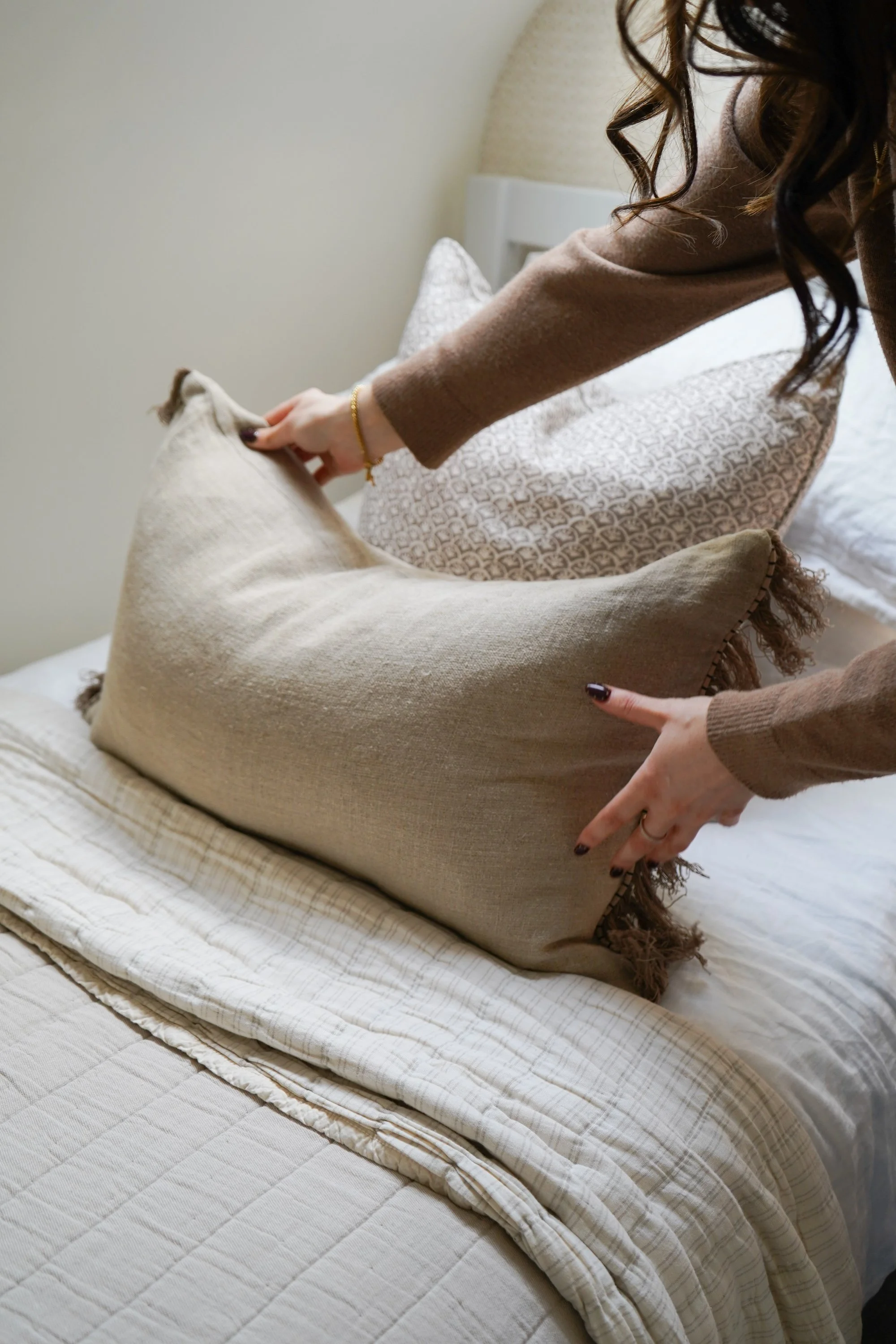 A person arranging decorative pillows on a bed in a bedroom with beige bedding and a beige headboard.