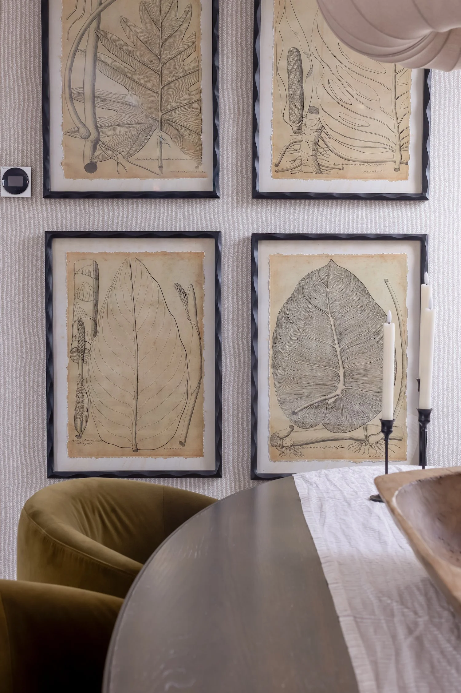 Wall art featuring botanical illustrations of leaves and plants, framed and displayed on a textured wall above a table with candles and a chair.