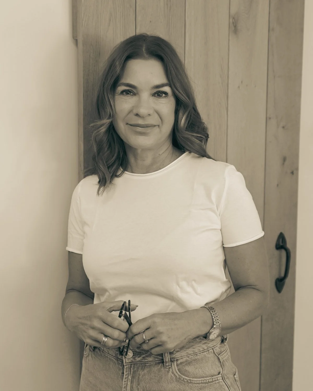 Black and white photo of a woman with shoulder-length wavy hair, wearing a white T-shirt and jeans, standing indoors next to a white wall and wooden closet door, holding glasses in her hand.