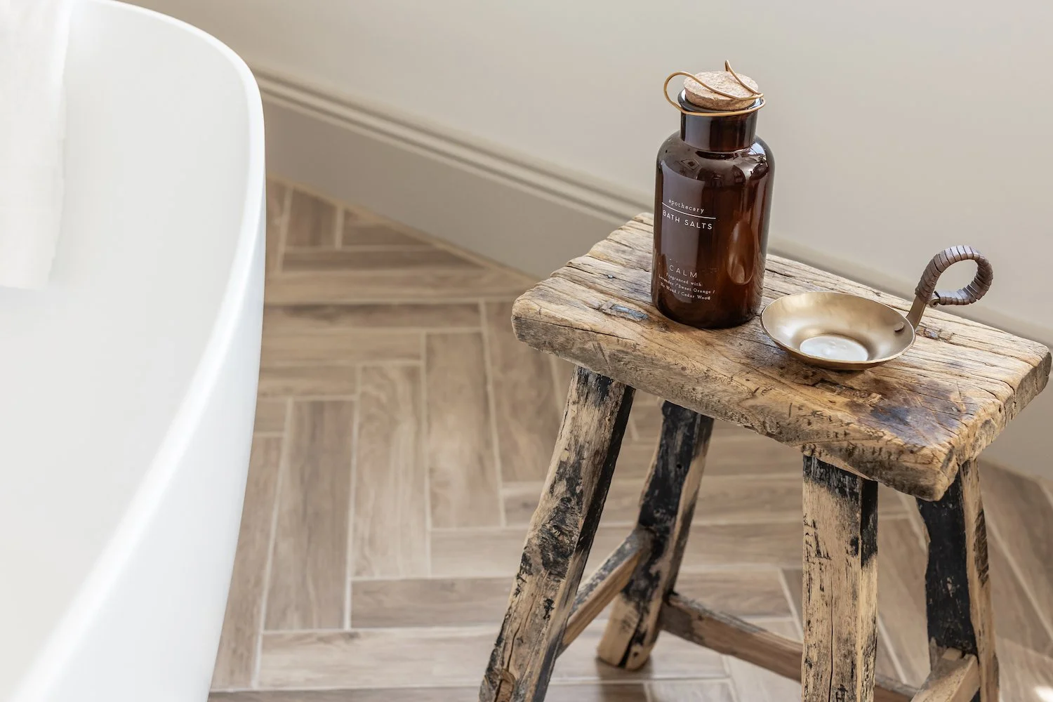 A rustic wooden stool with blackened legs, placed on a tiled wooden floor, holding a brown glass bottle of bath salts labeled 'Calm' and a small metallic dish with a curved handle.