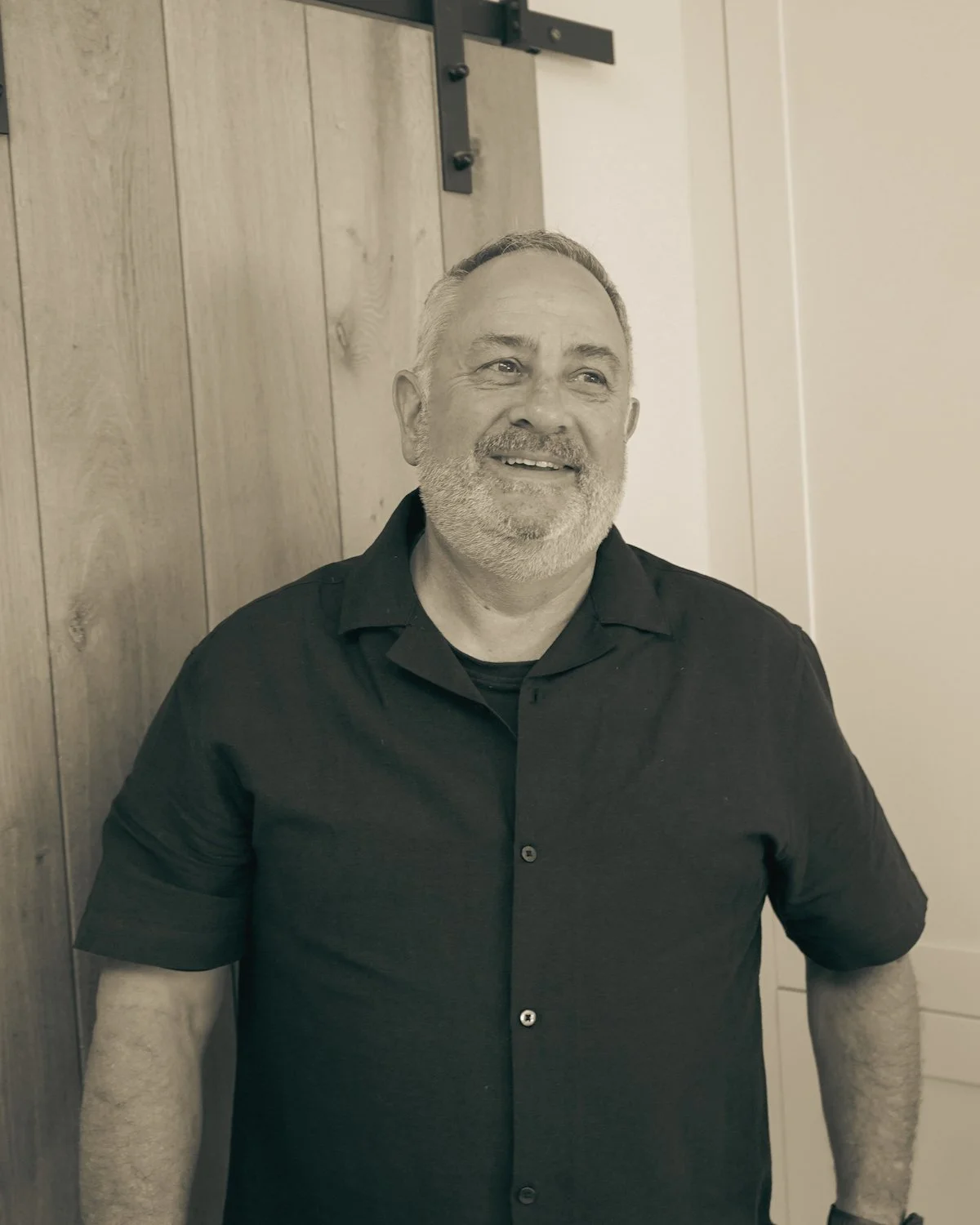 A smiling older man with gray hair and a beard standing in a kitchen, leaning against a wooden wall with cabinetry visible behind him.