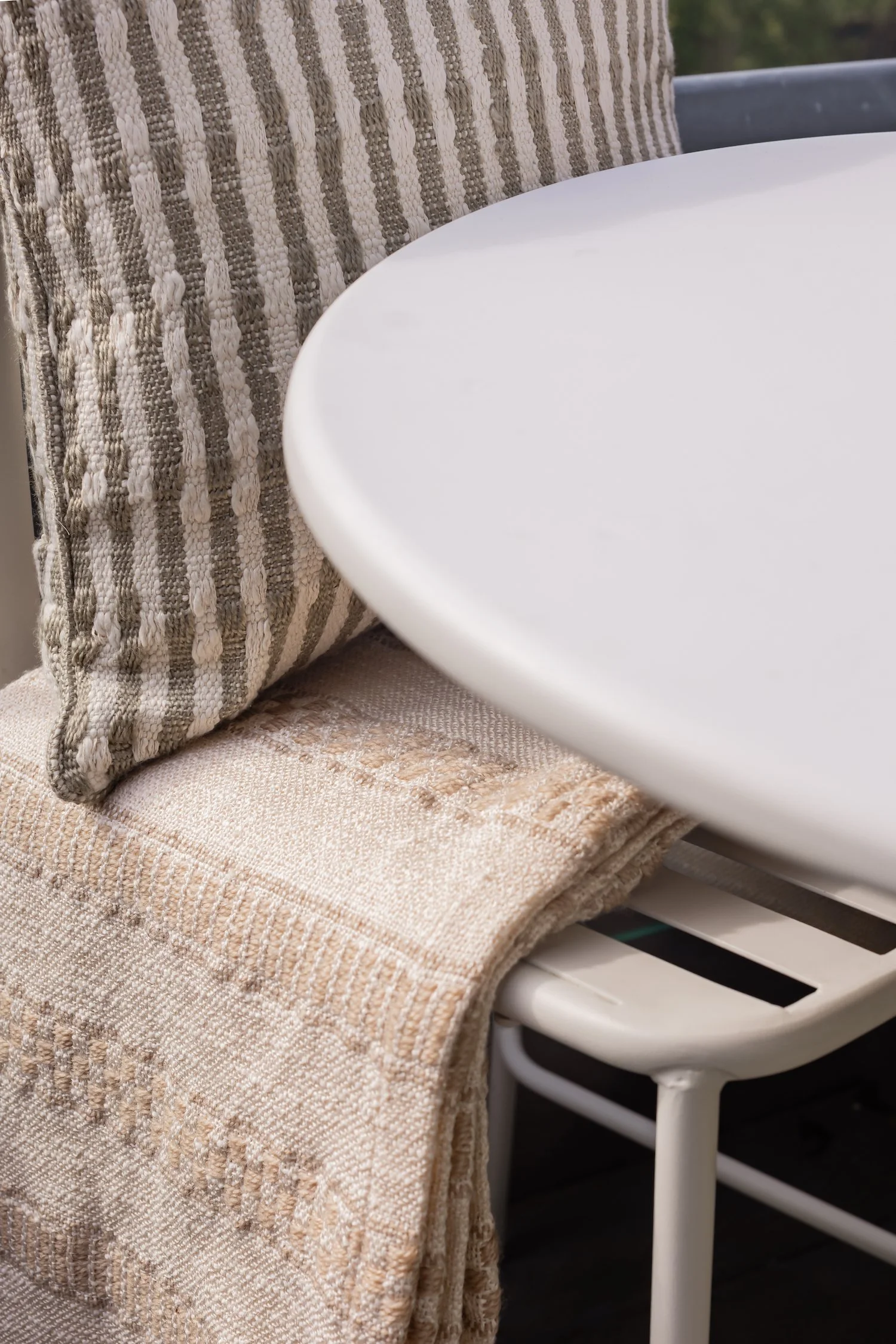 Close-up of a beige woven blanket and a textured beige towel draped over a white chair, with part of a white round table visible.