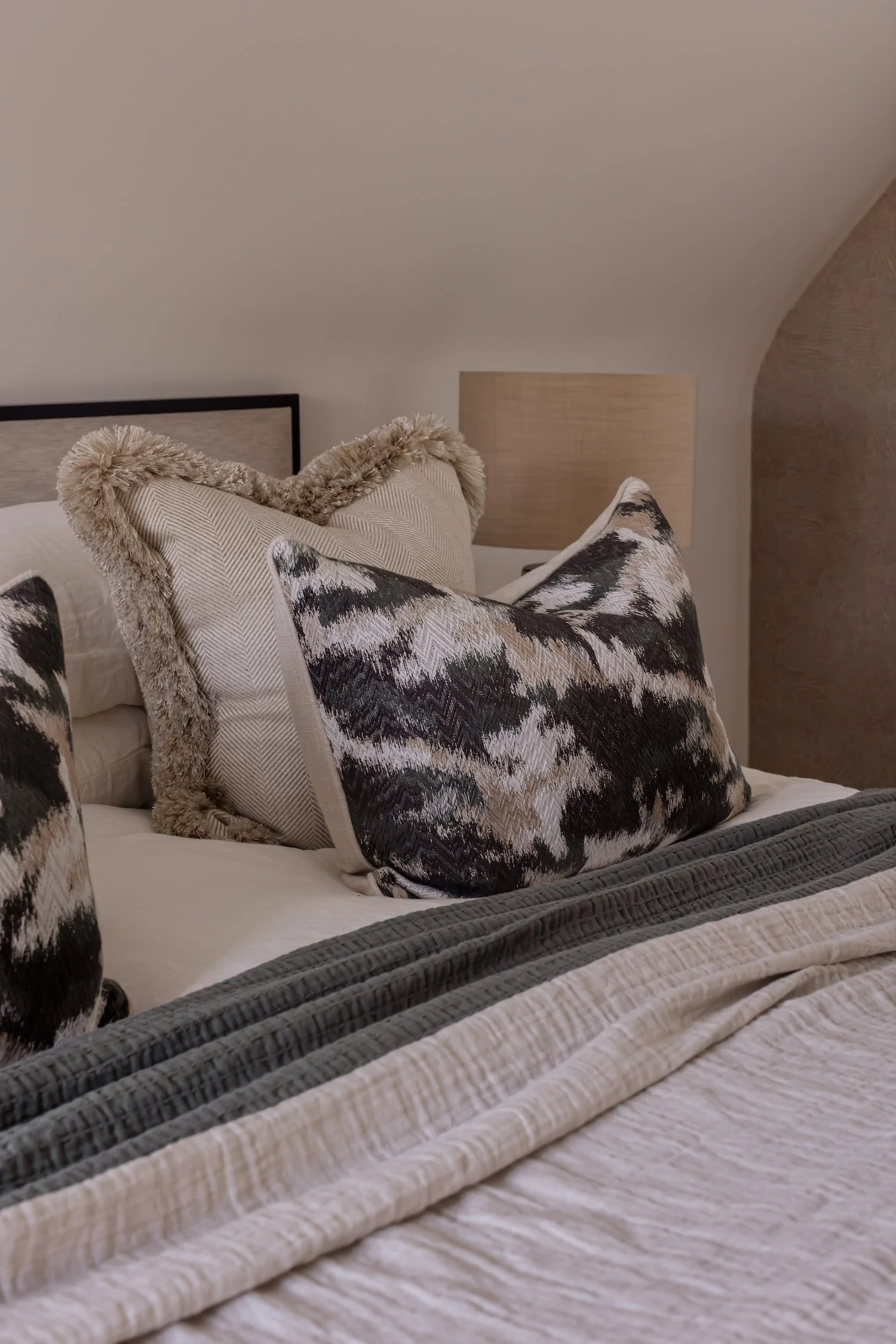 Close-up of a bed with decorative pillows featuring black and white abstract patterns and a beige throw blanket, in a bedroom with neutral-colored decor.
