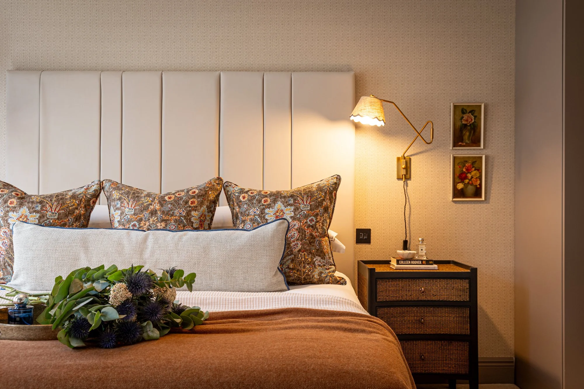 A bedroom with a white padded headboard, floral-patterned pillows, a beige wall with two framed floral paintings, a wall-mounted reading lamp, a small black nightstand with books and decorative items, and a flower arrangement on the bed.