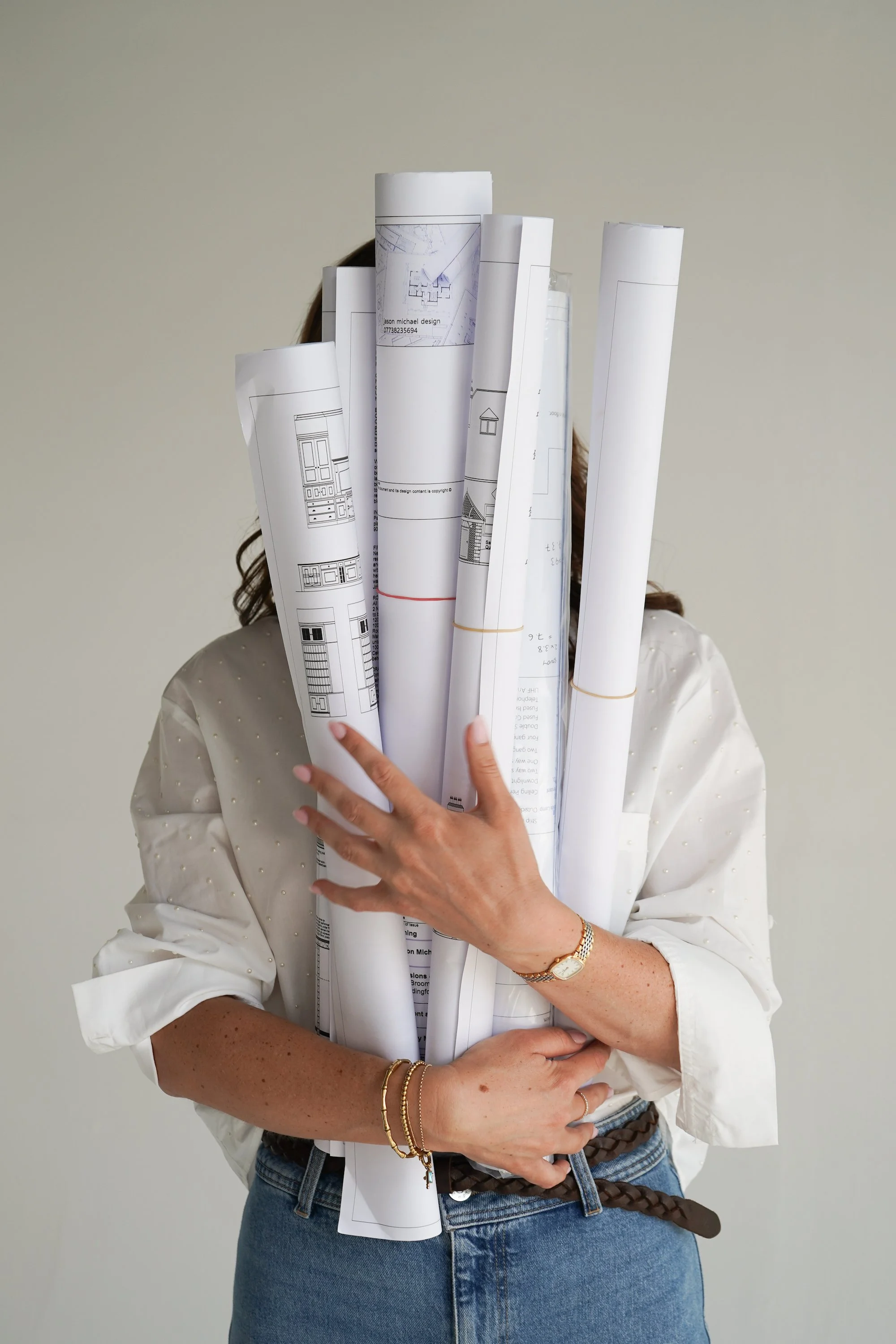 Person holding several rolls of architectural blueprints in front of their face, wearing a white blouse, blue jeans, and gold jewelry, with a plain background.