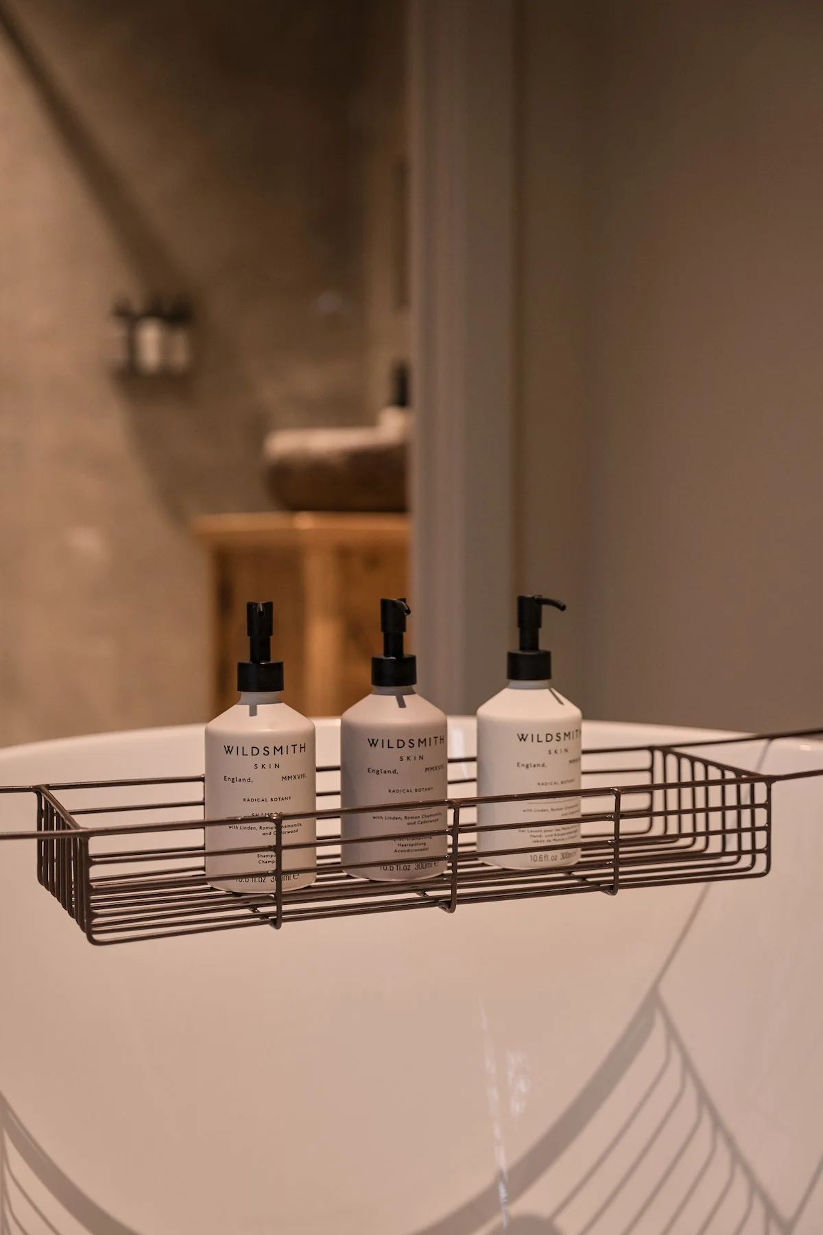 Three bottles of Wildsmith skin care products in a metal shower caddy, mounted on a bathroom wall, with a blurry background of a sink and countertop.