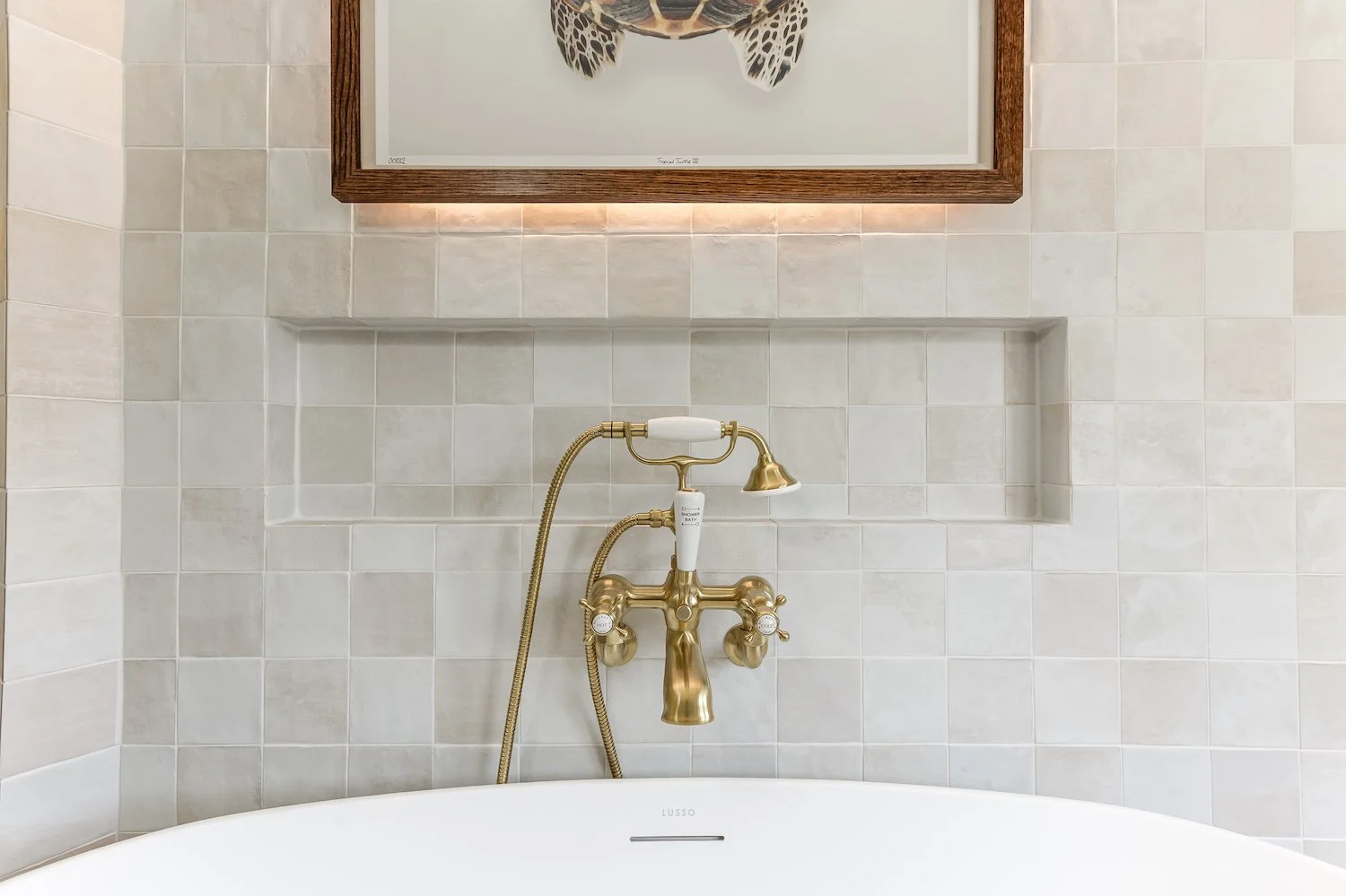 A bathroom with a white bathtub, a brass vintage-style faucet with a handheld showerhead, and a framed sea turtle artwork above the bathtub. The wall is tiled with beige tiles, and there is a recessed shelf in the wall.