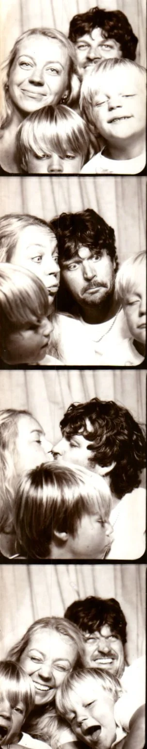 Four black and white photo strips featuring a woman with long hair, and a man with curly hair, with two children with light hair. In the first strip, they are smiling and close together. In the second, the woman and man look surprised or amused. In the third, they are about to kiss, and in the fourth, they are laughing and smiling together.