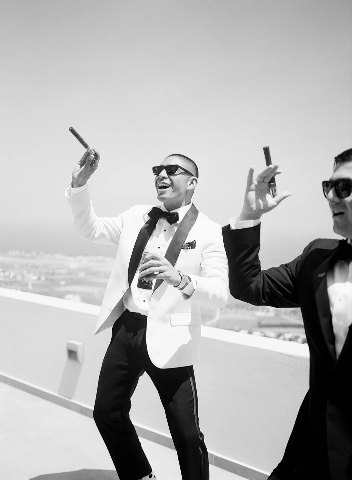 Two men in tuxedos celebrating, holding cigars and drinks, enjoying sunny weather outdoors, with a cityscape in the background.