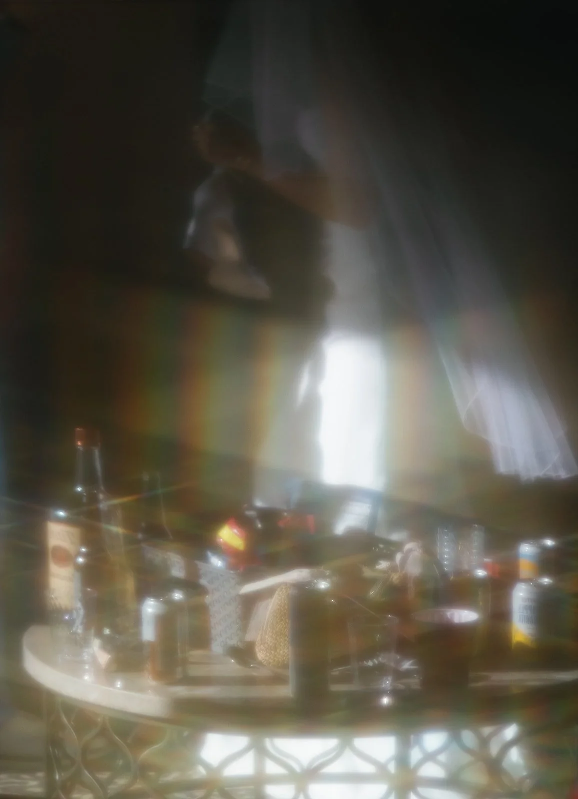 Blurred image showing a person behind a semi-transparent surface, with a table of various drinks and bottles in front, possibly in a bar or restaurant setting.