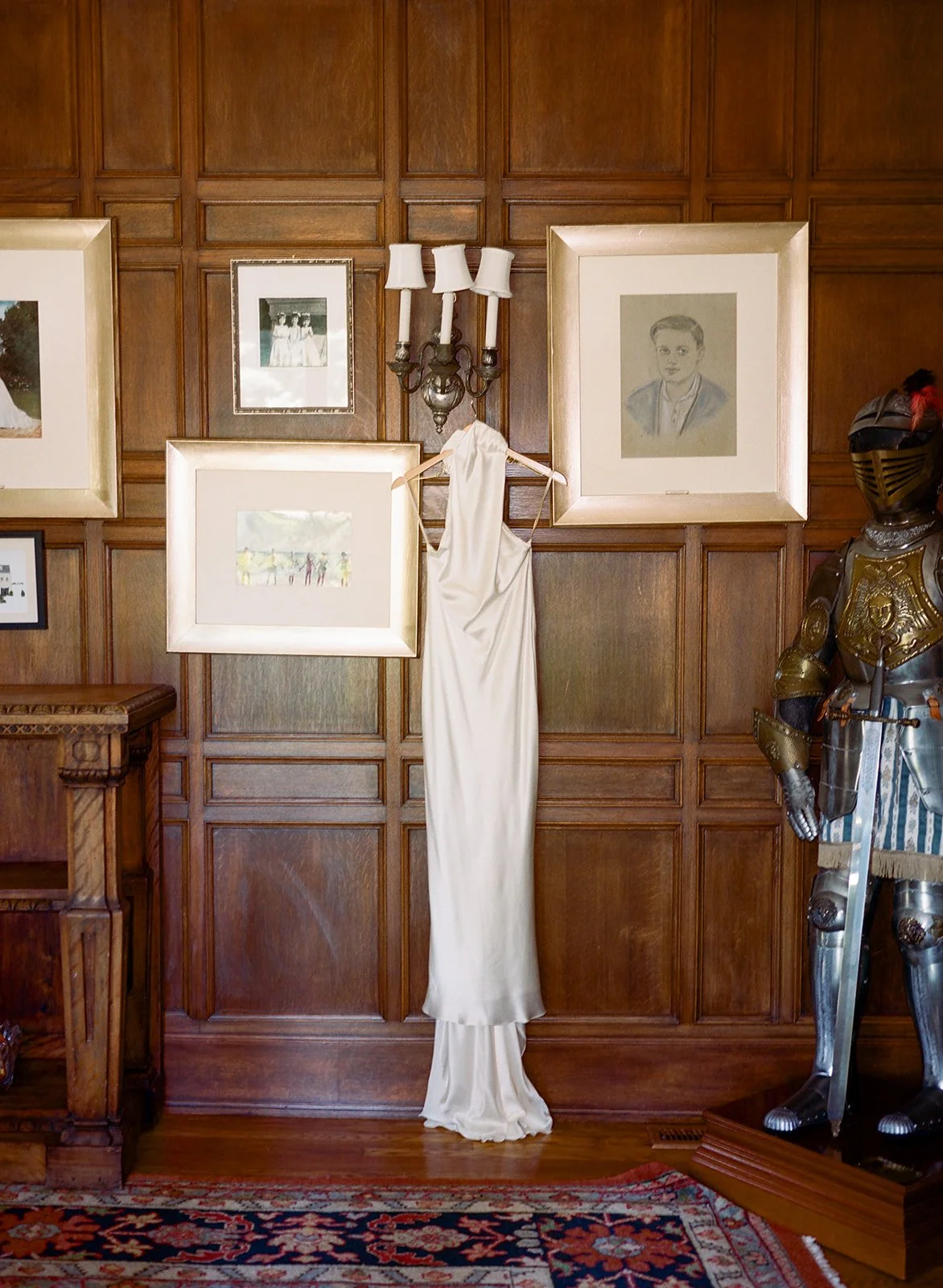 Wood-paneled wall decorated with framed artwork, a sconce light, a long white dress hanging, and a suit of armor.