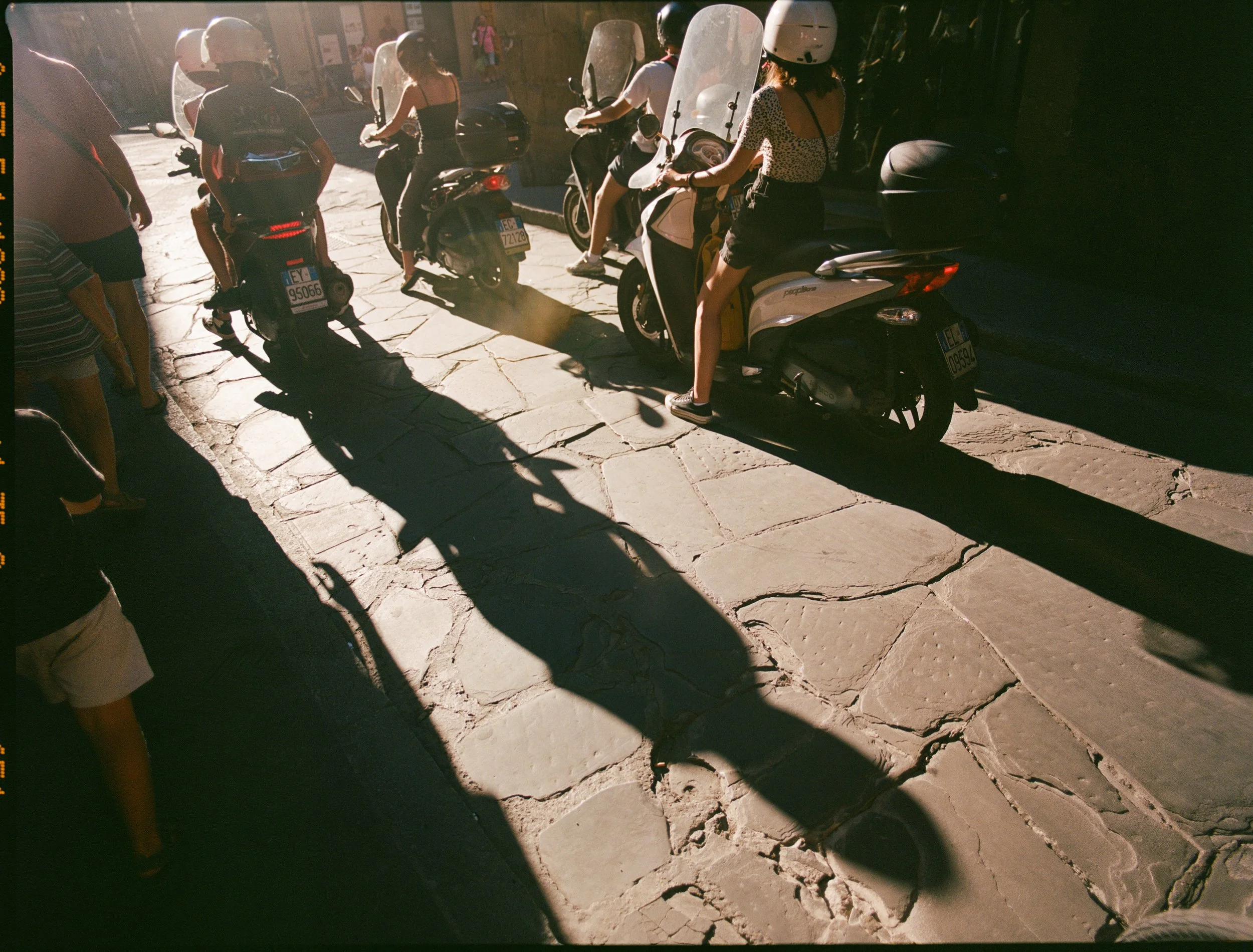 People with helmets sitting and standing near scooters on a sunny sidewalk.