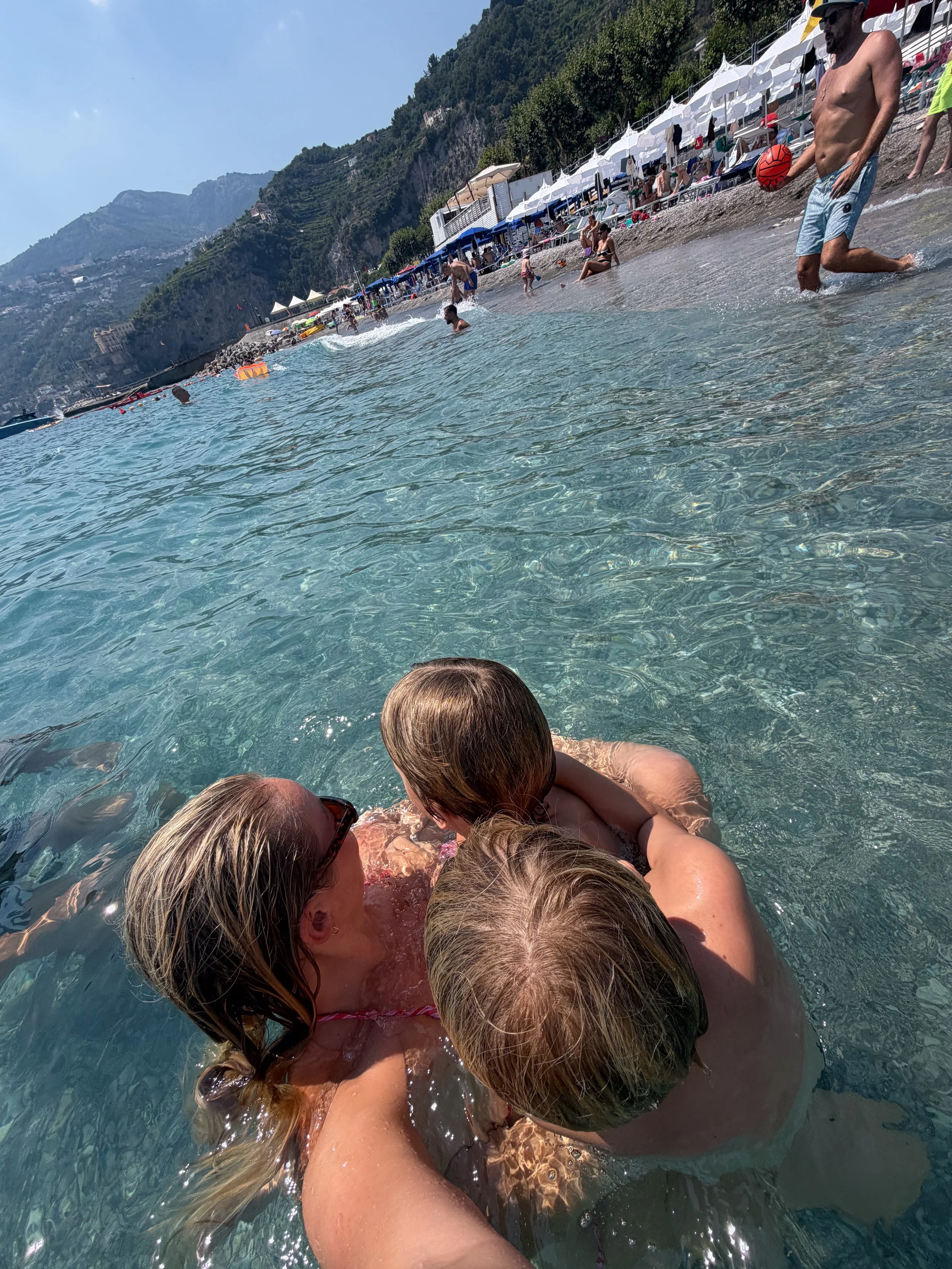 People swimming and relaxing at a beach with umbrellas, overlooking mountains.