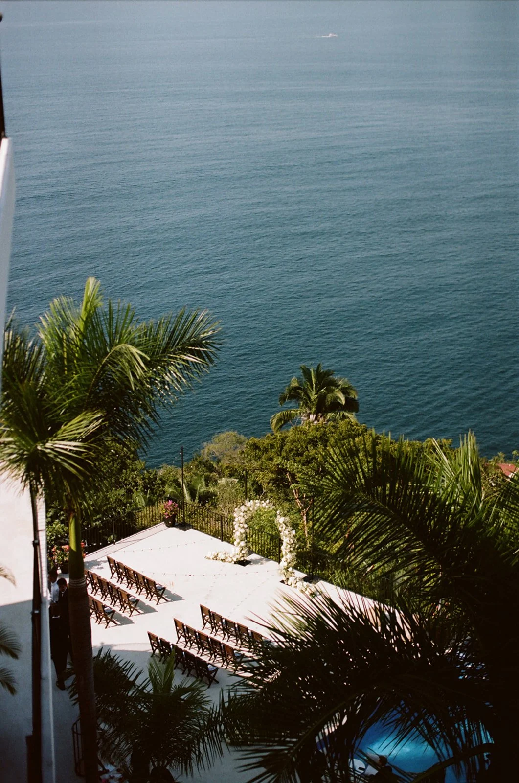Outdoor wedding ceremony setup with chairs in front of a floral arch, overlooking a body of water, surrounded by palm trees and lush greenery.