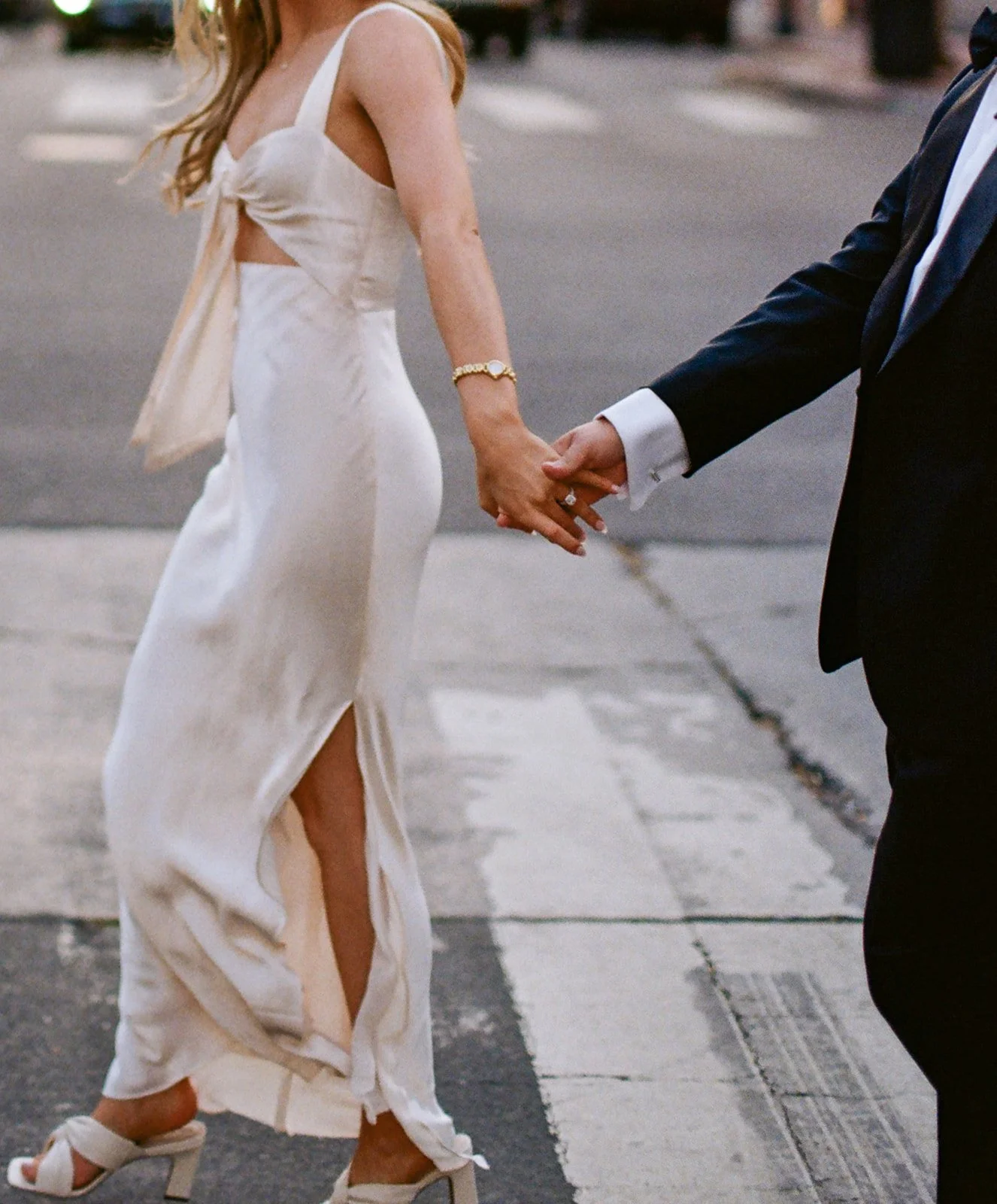 A woman in a white dress and high heels holding hands with a man in a suit, walking together on a street.