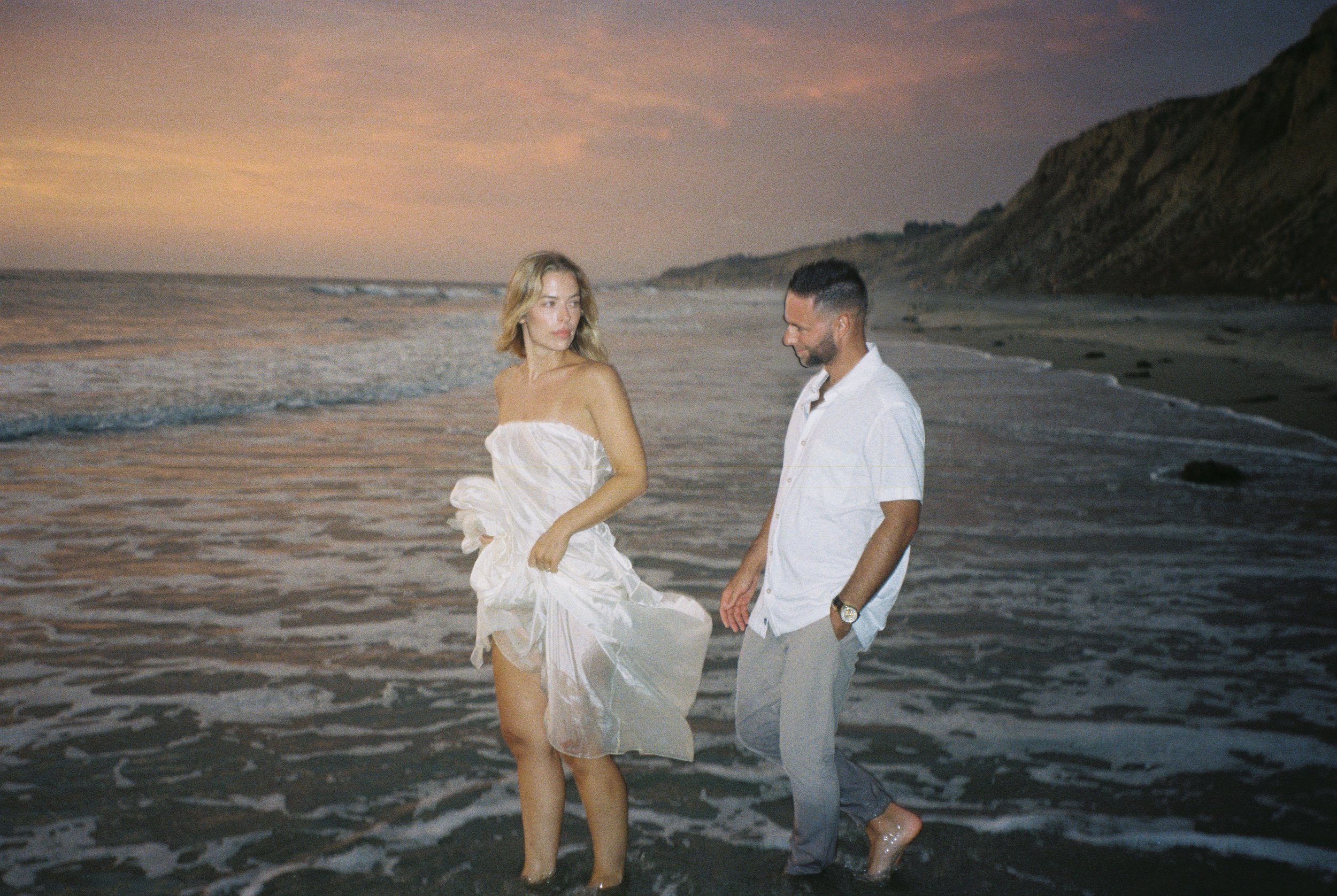 A woman in a white dress and a man in a white shirt walking along the shoreline at sunset, with cliffs in the background.