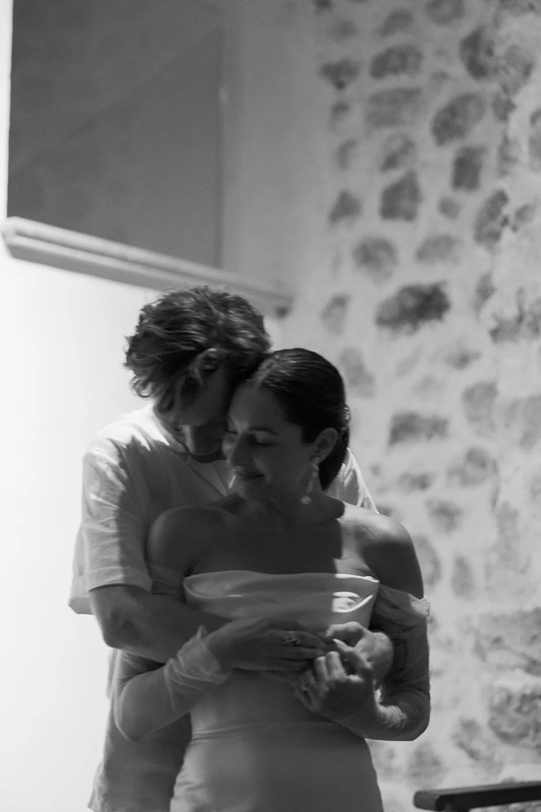 A black and white photo of a couple embracing, with the man leaning down to kiss the woman's forehead. The woman is smiling and looking away. They are standing indoors near a stone wall.