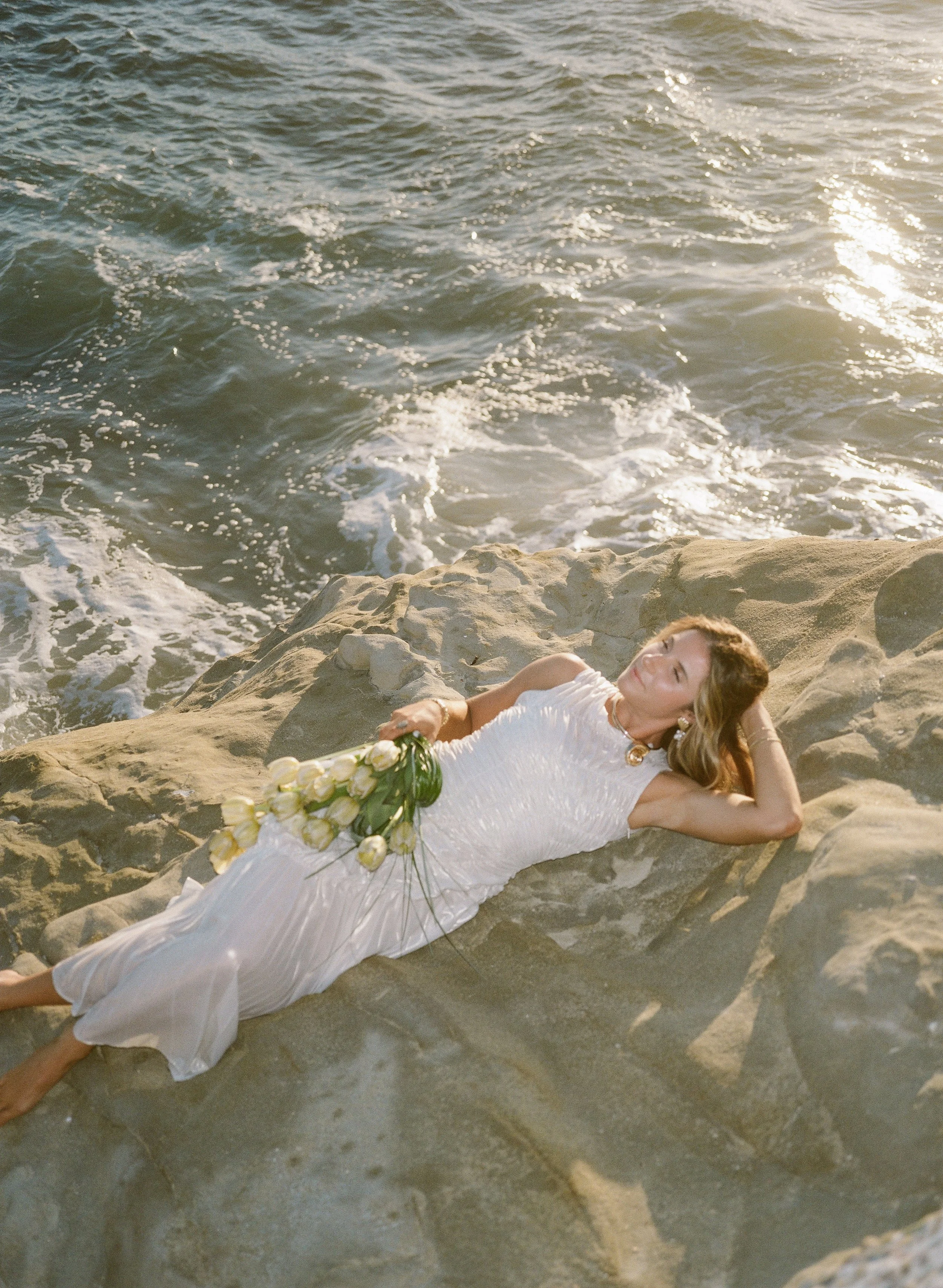 Woman in white dress lying on rocks by the seaside with a bouquet of white tulips.