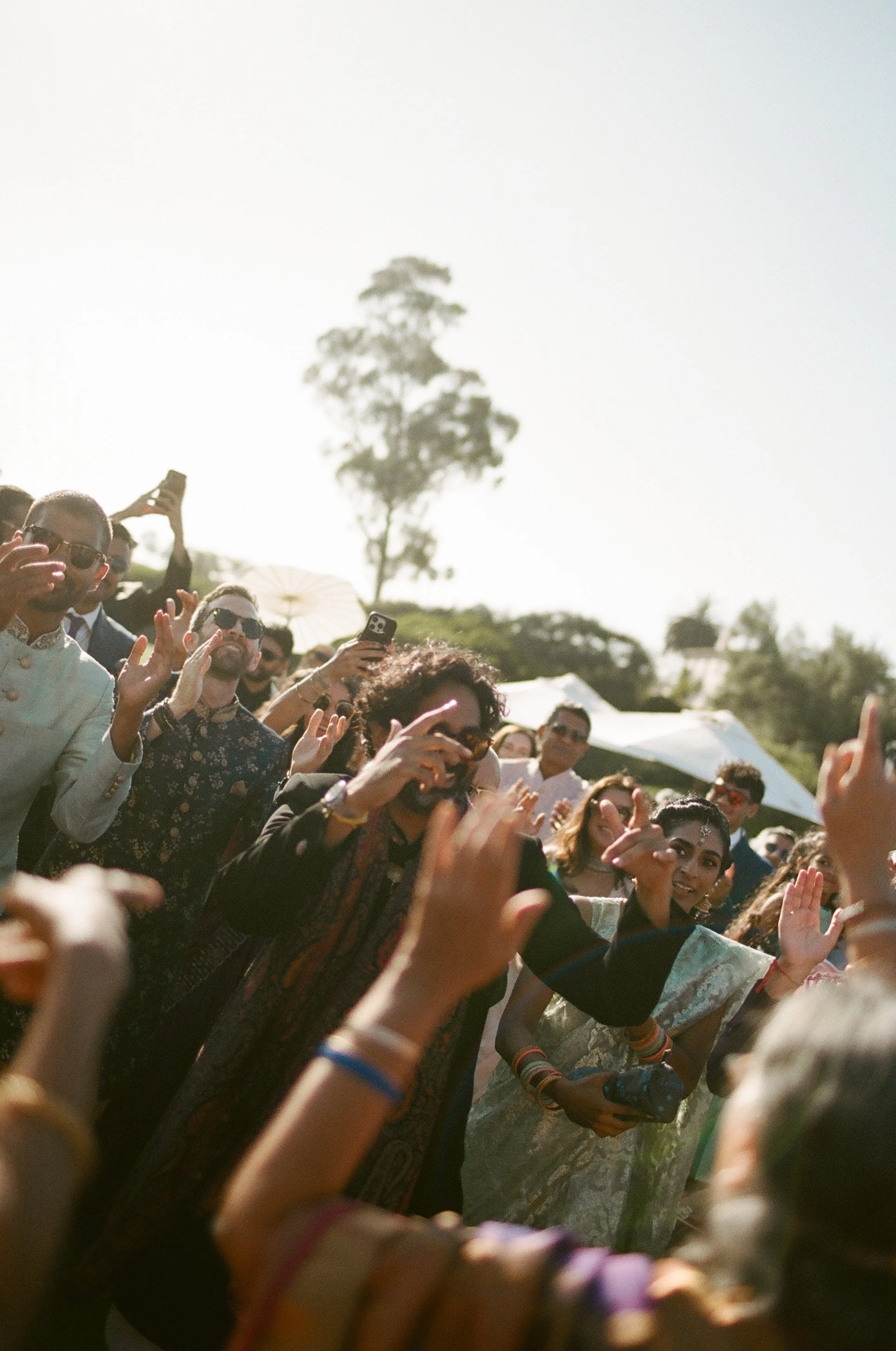 Crowd of people attending an outdoor event, some taking photos or videos with their smartphones, with trees and tents in the background under a clear sky.