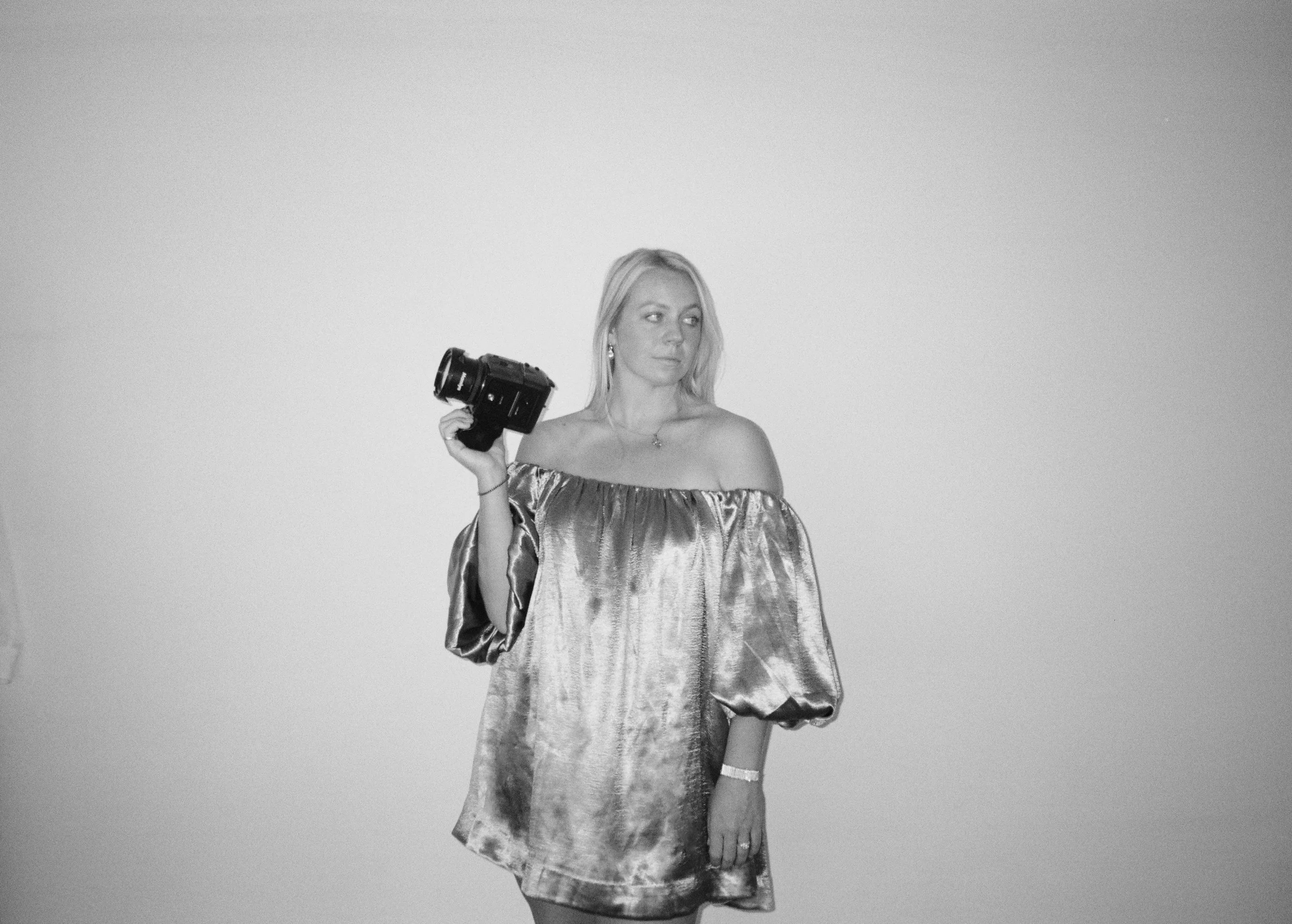 A woman stands against a plain background, holding a camera in her right hand, wearing an off-the-shoulder dress with puffy sleeves.