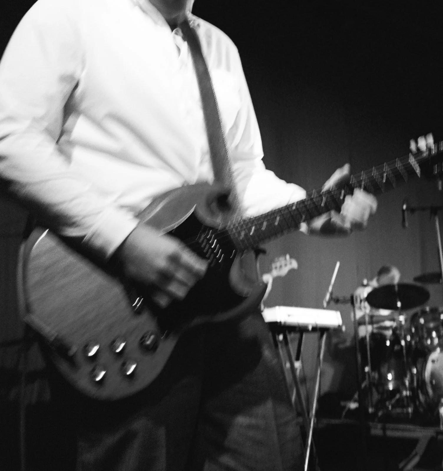 Black and white image of a person wearing a white shirt playing an electric guitar. There are other musical instruments and equipment visible in the background, indicating a music performance or rehearsal setting.