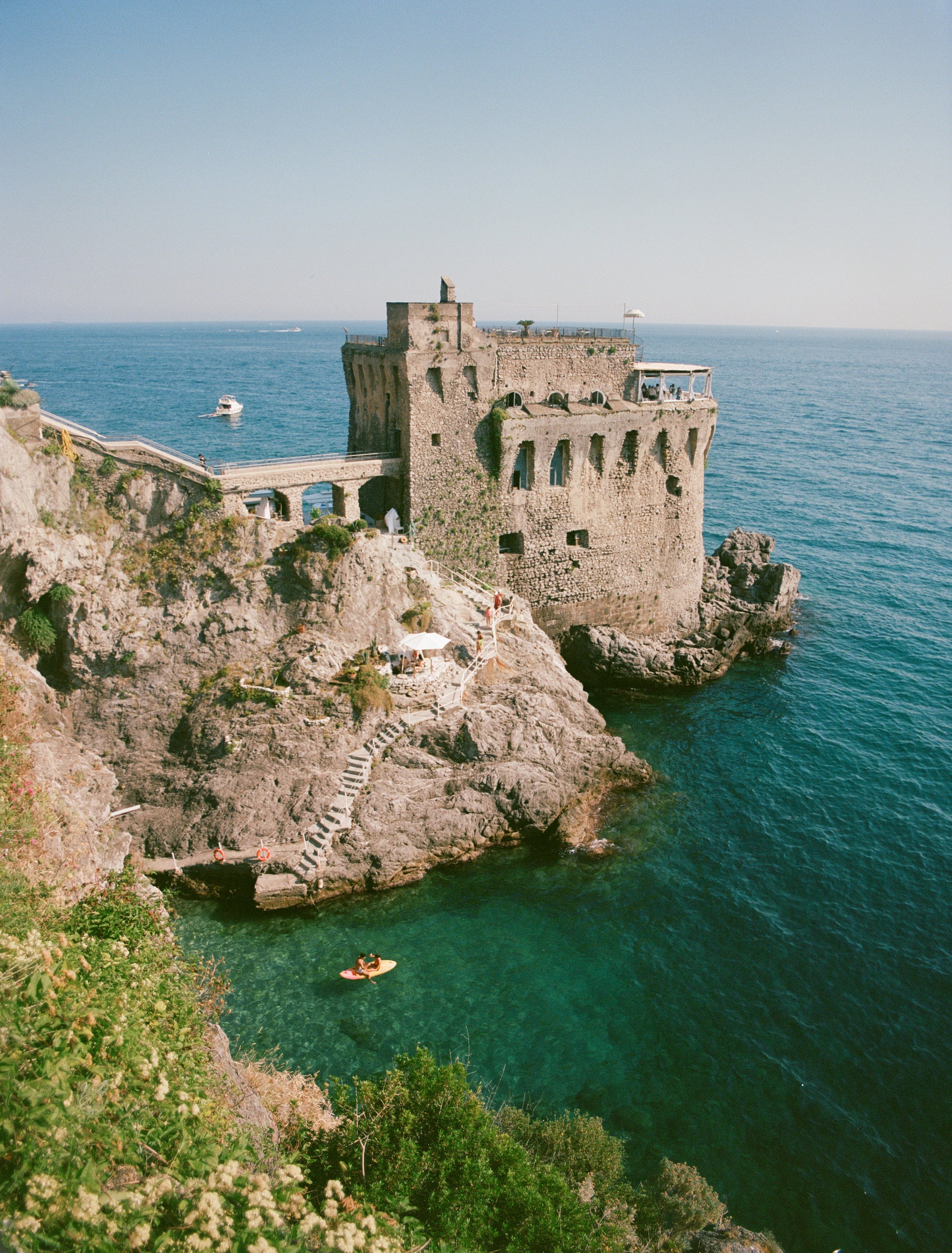 A historic stone castle on a rocky coastline overlooking the ocean. The castle has arched windows and a flat roof with a small rooftop structure and a white umbrella. A staircase is built into the cliffside leading down to the water, where two people enjoy paddleboarding in the greenish-blue sea. Green shrubbery and white flowers are in the foreground, with a clear sky above.