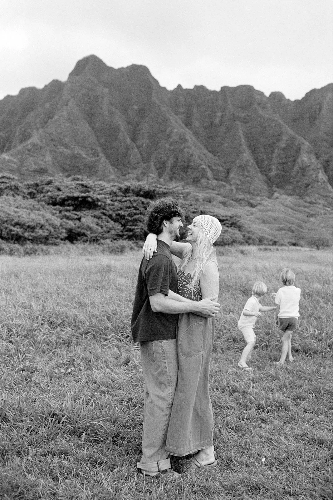 A couple stands close together, looking into each other's eyes, smiling at a scenic mountain backdrop. Two children are playing in the grass behind them.