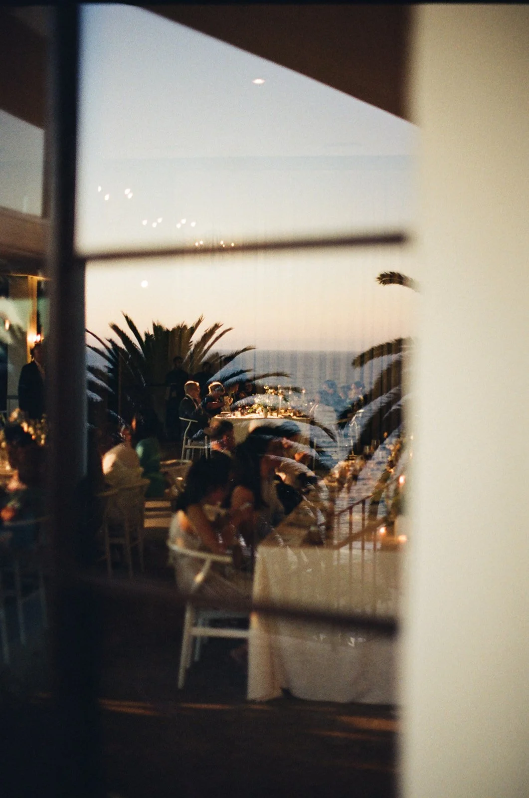 A reflection of a dining event outdoors at sunset, seen through a window, with tables, people, palm trees, and the ocean in the background.
