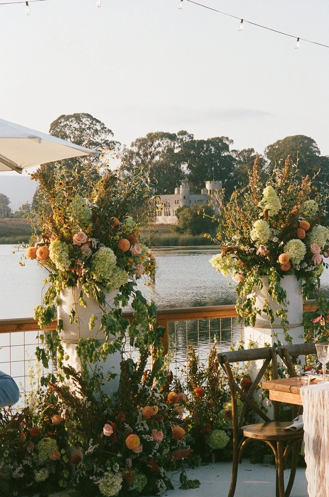 Decorative floral arrangements on a balcony overlooking a lake with a castle in the distance during sunset.