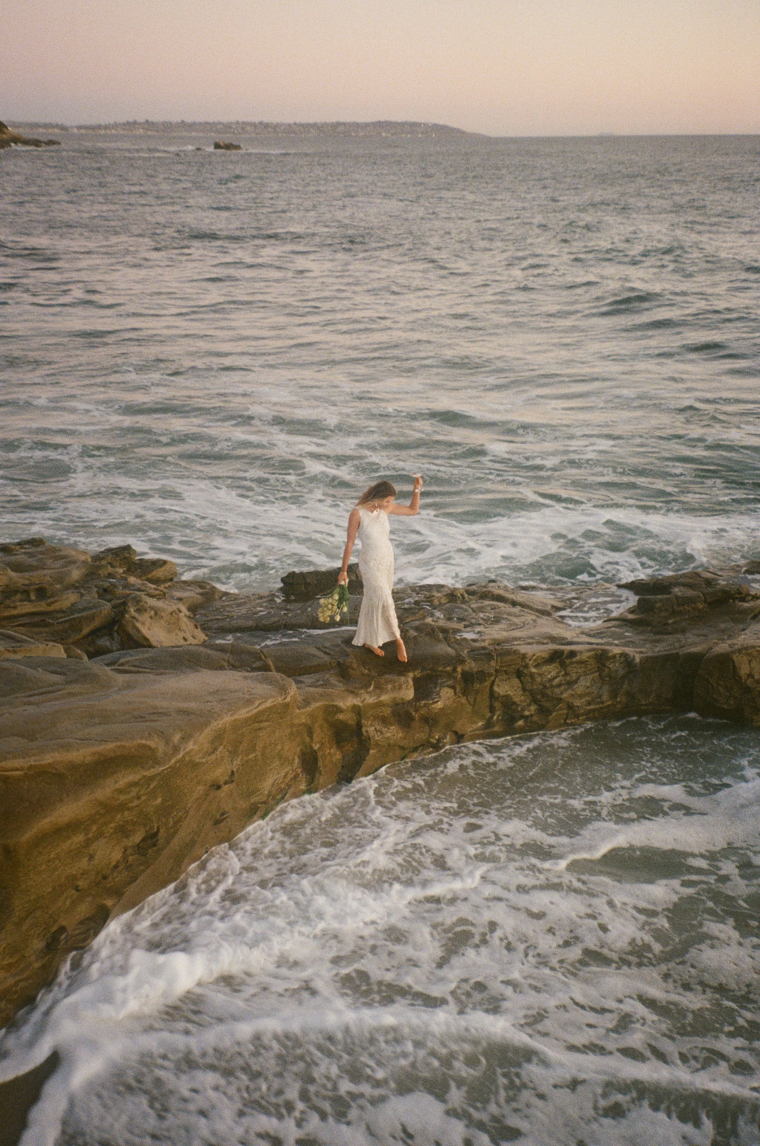 A woman in a white dress walking on rocks by the ocean at sunset, holding a bouquet of flowers.